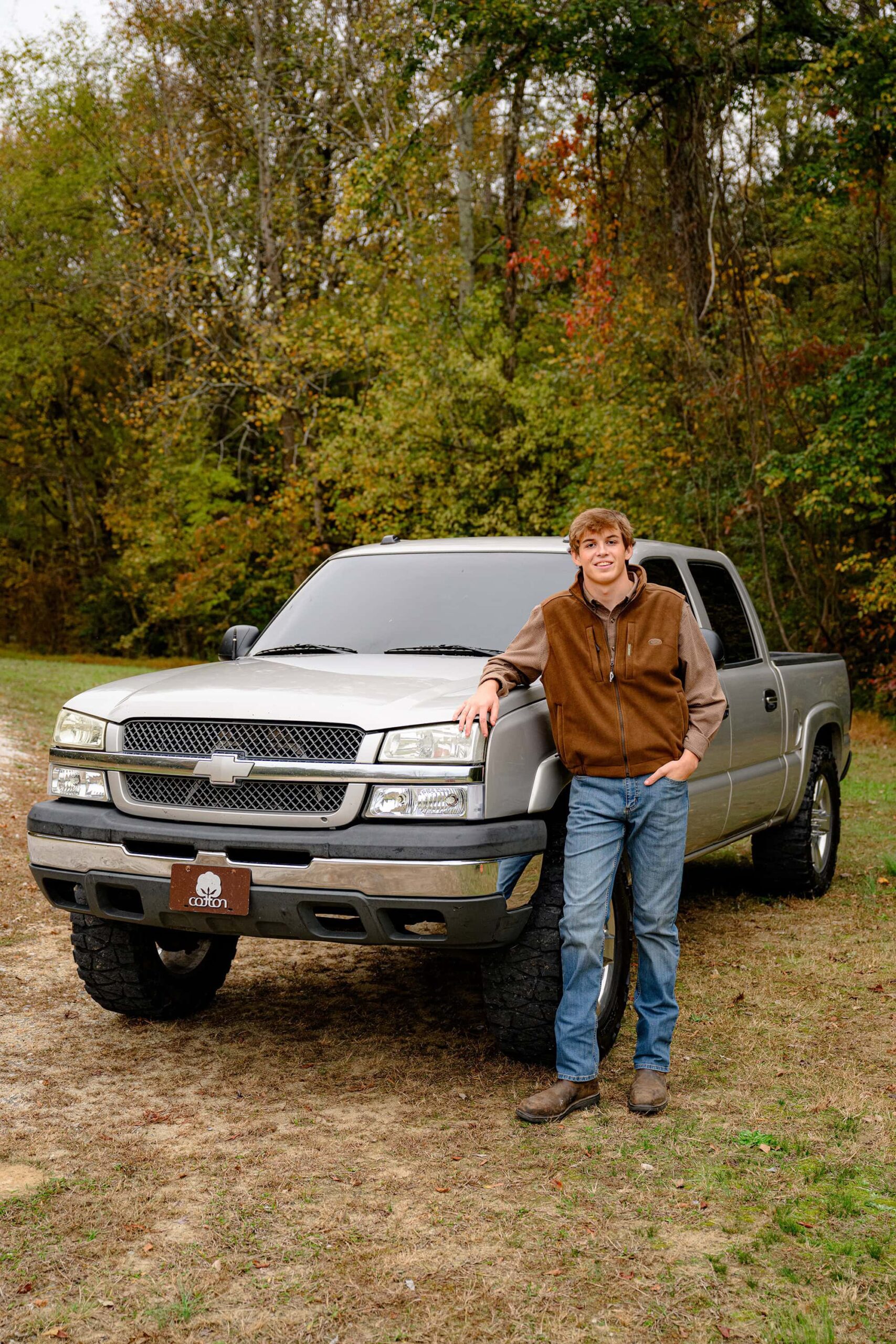 A senior boy from Lancaster, SC, wearing a brown vest, plaid shirt, and jeans, leaning against his truck with the truck bed open, surrounded by a scenic rural landscape.