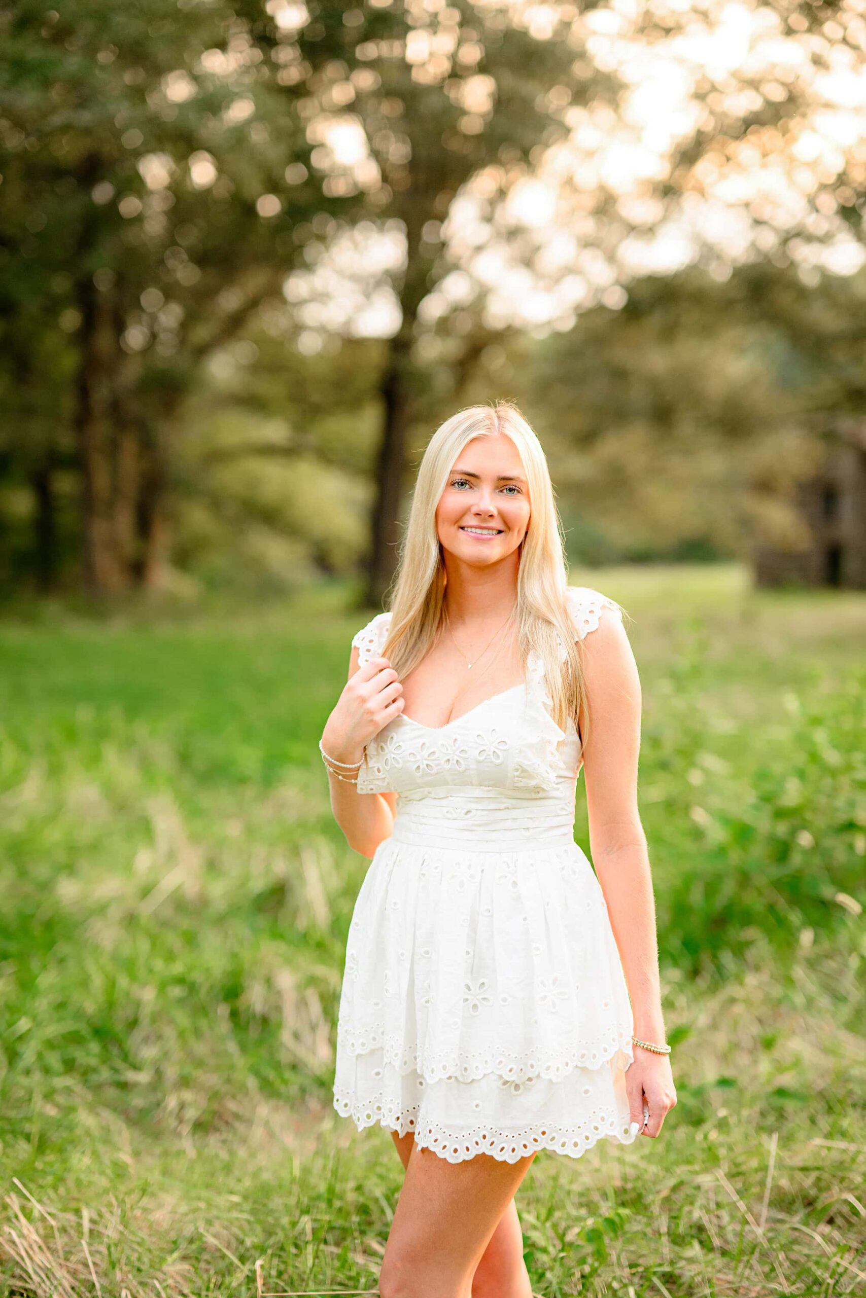 A senior with long blond hair and green eyes from Cuthbertson High School, wearing a creamish-white baby doll dress, standing gracefully in a lush green field, with soft sunlight illuminating the scene.