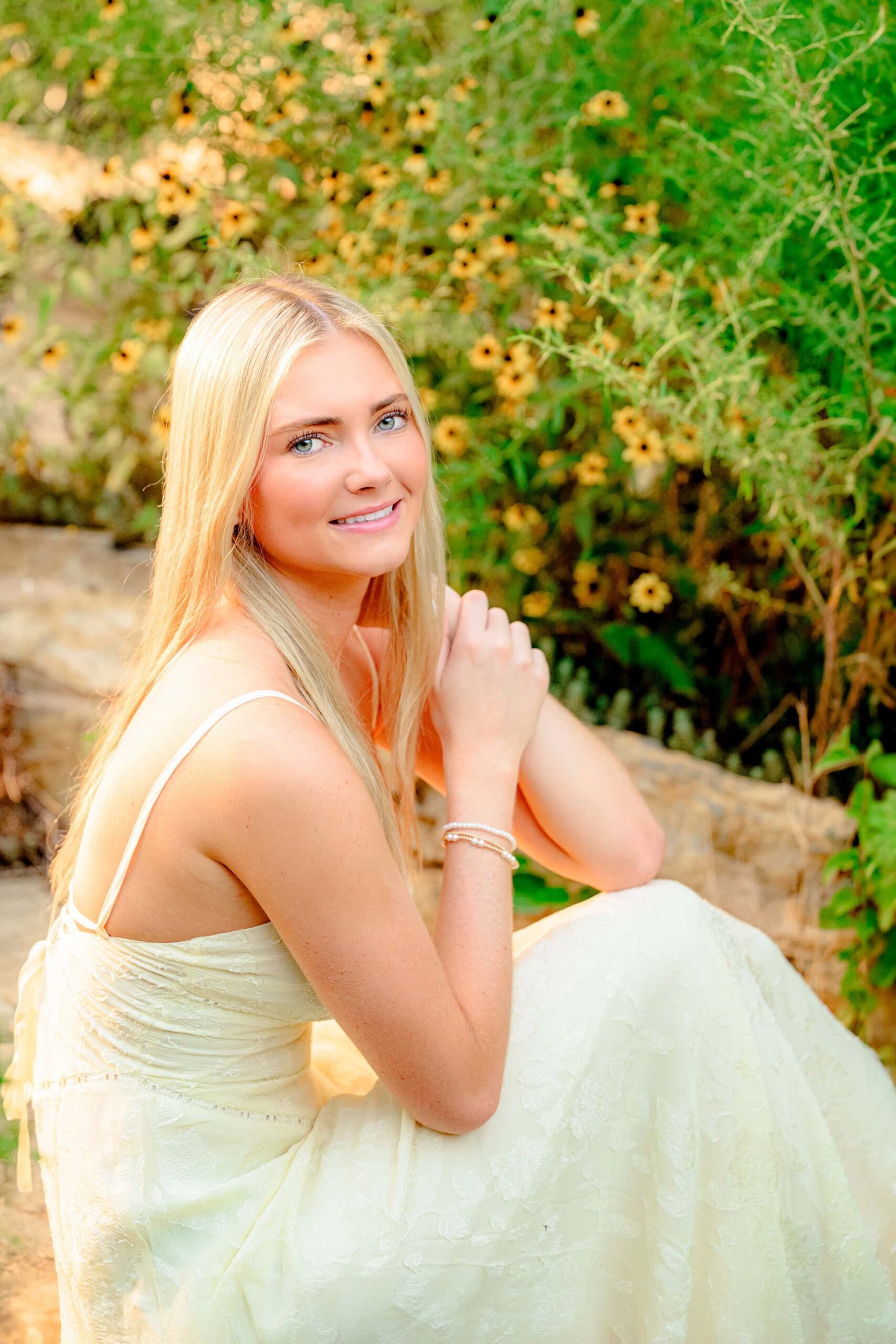 A senior from Cuthbertson High School with long blond hair and green eyes, wearing a greenish-yellow dress, sitting on stone steps surrounded by yellow flowers, exuding a relaxed and natural vibe.