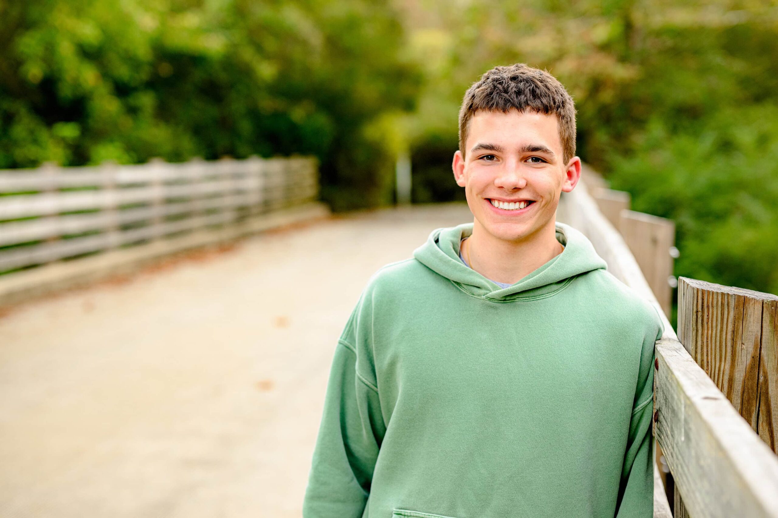 A C.H.S. grad with short dark hair, wearing a green shirt, leaning on the rail of a bridge with vibrant fall colors in the background, creating a thoughtful and calm vibe.