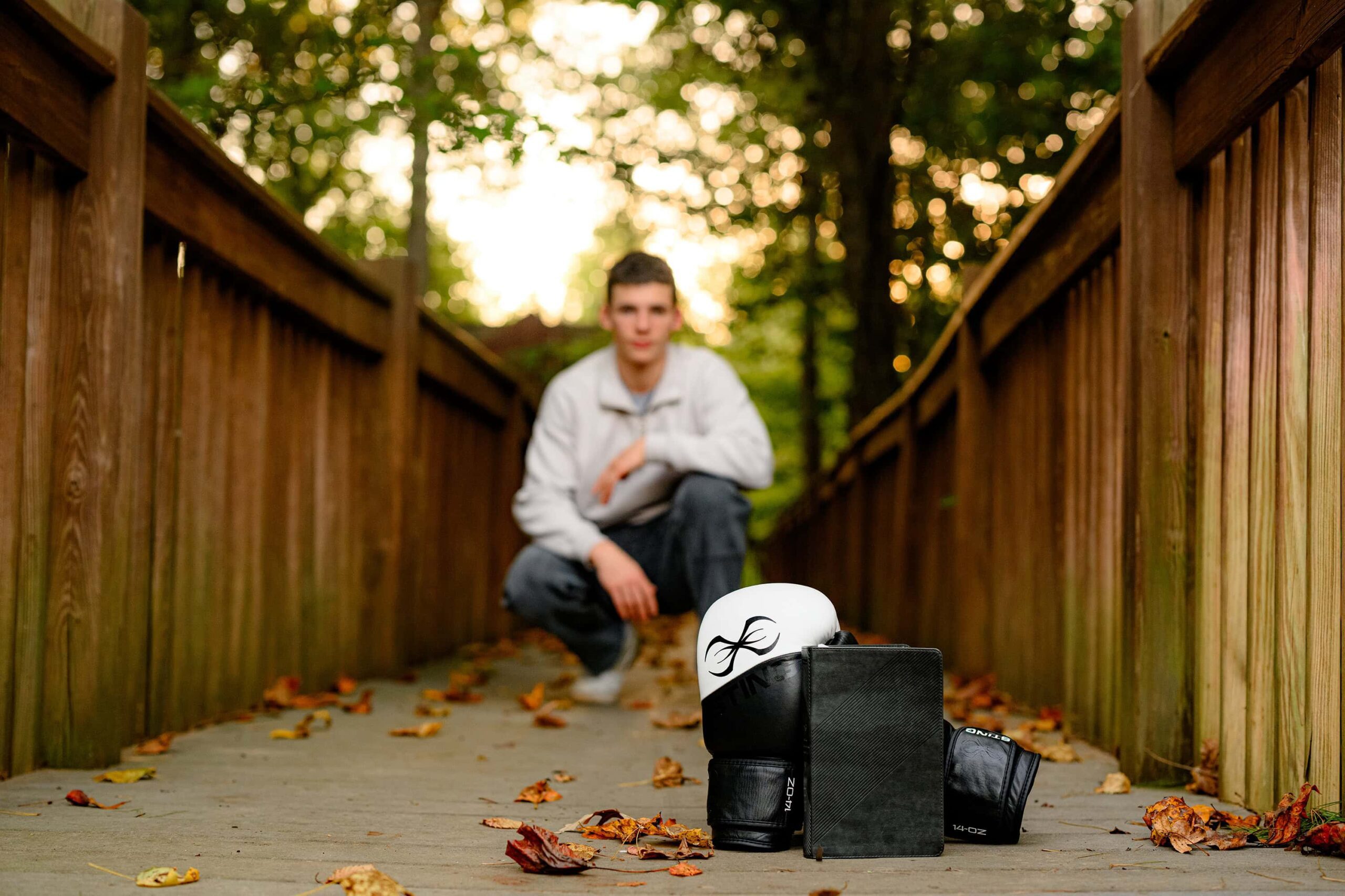 The same grad in a cream shirt and dark pants, holding his Bible with boxing gloves placed in the foreground, framed by double railings on each side, capturing a meaningful, personal moment.