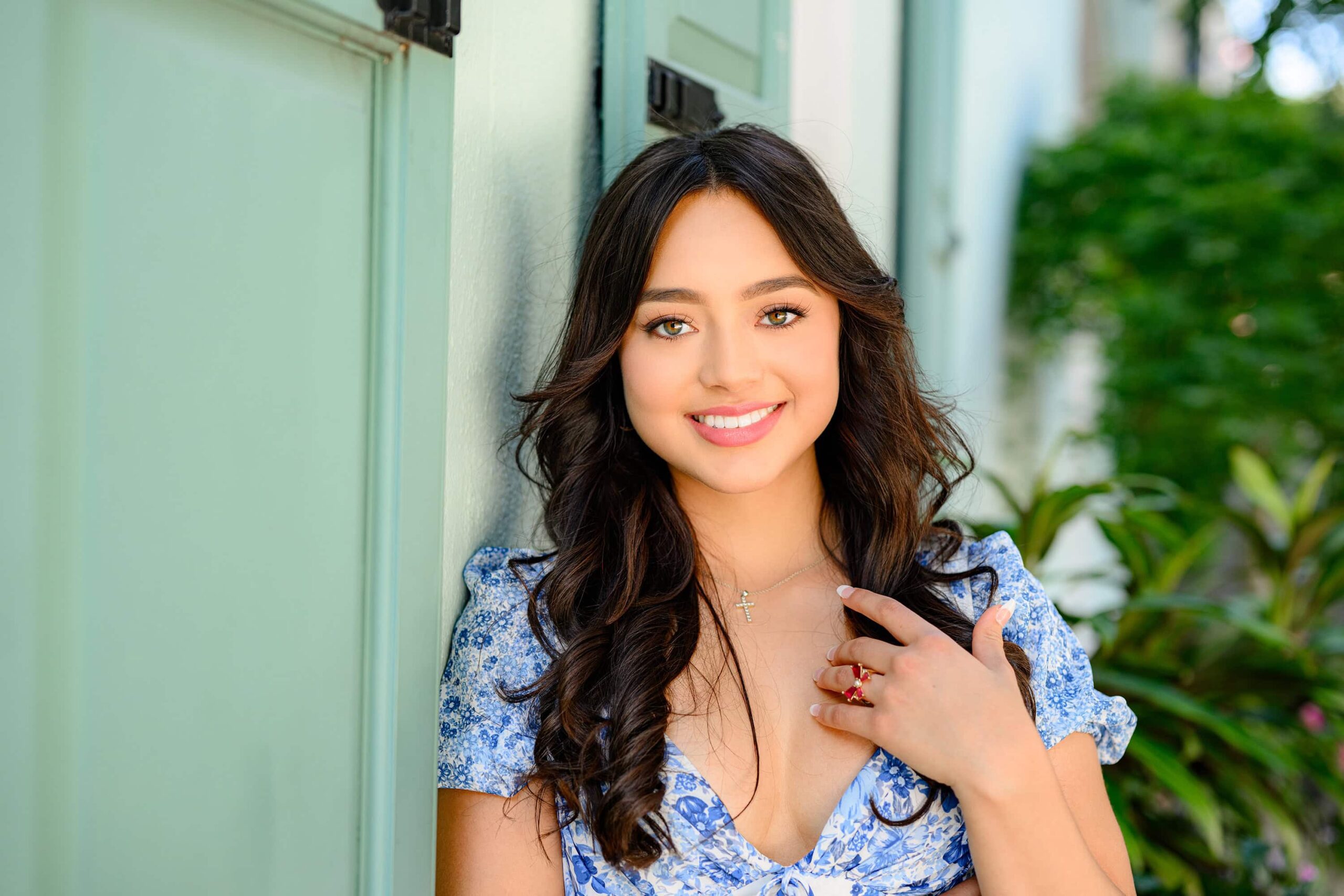 A Union County, NC grad with long dark hair and bright green eyes, wearing a blue print dress, leaning against a vibrant green wall in Charleston, SC, creating a stylish and timeless moment.