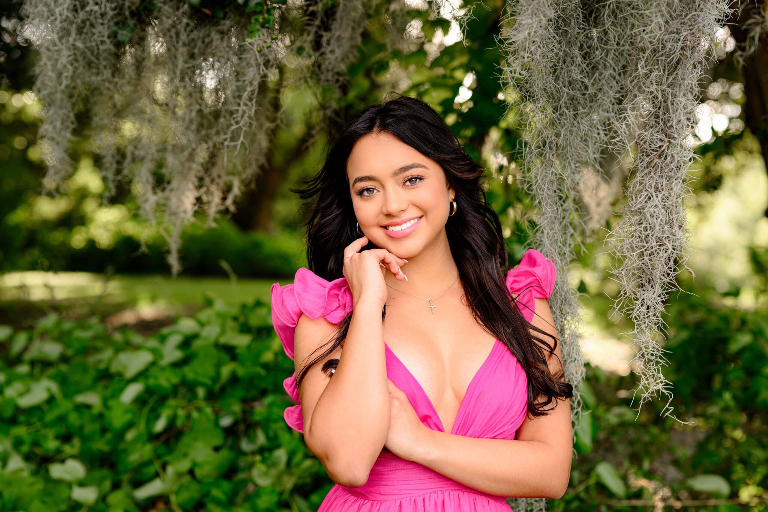 A grad with long dark hair and green eyes, wearing a bright pink dress, poses in a Charleston, SC park. Her hair blows in the wind as she folds her arms, gently touching her cheek, with the iconic Spanish moss creating a soft, timeless backdrop.
