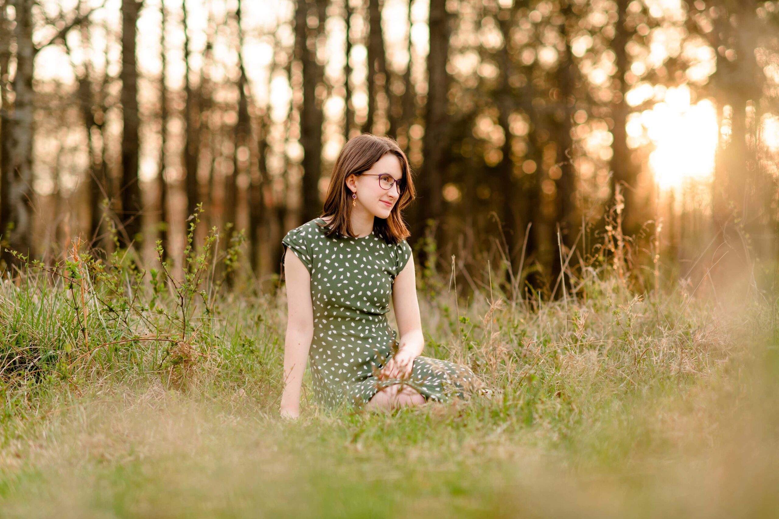 sitting in the tall grass a senior looks off in the distance with the golden light shining on her wearing a green dress