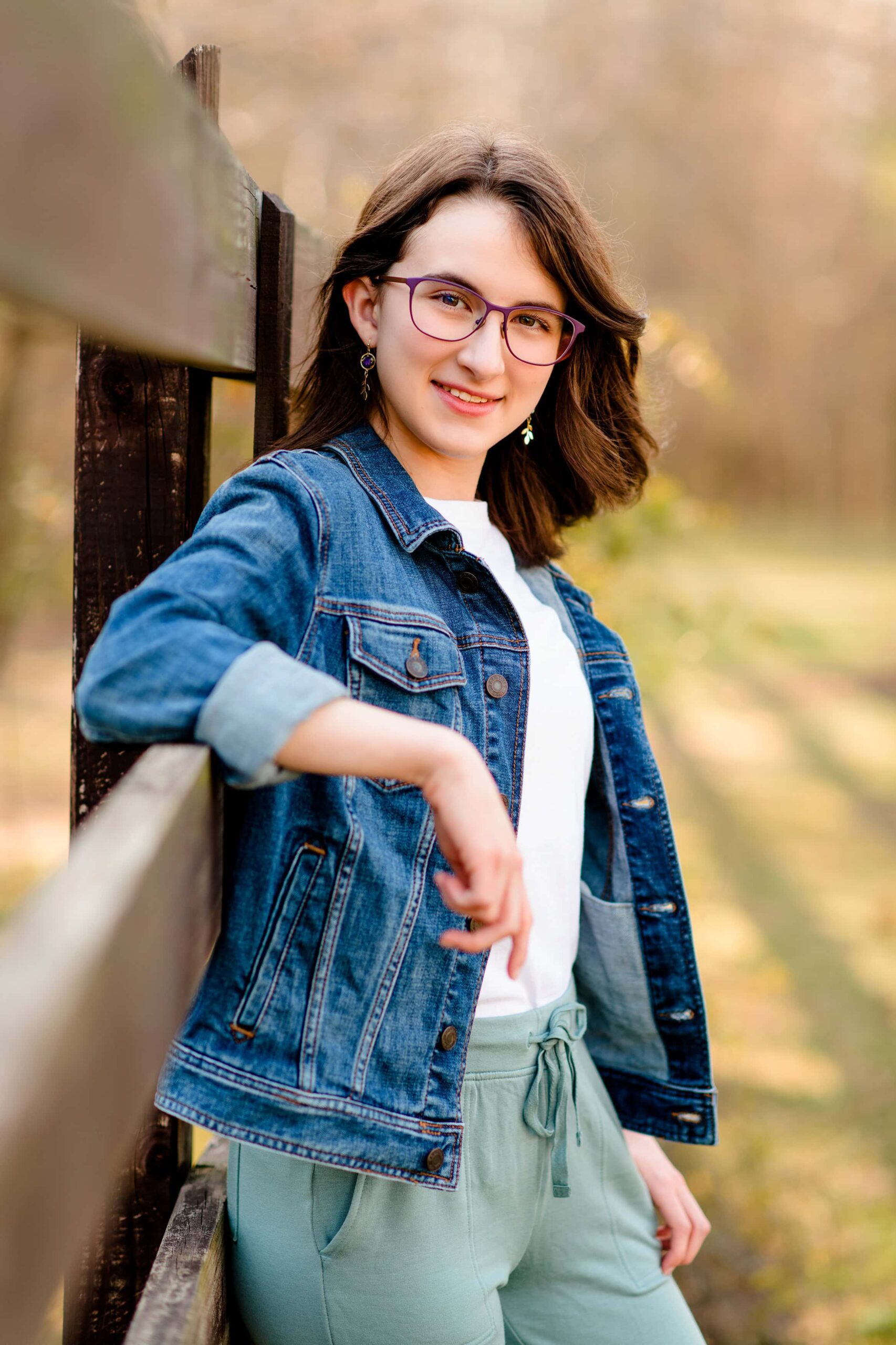 dark hair senior girl leans against a fence wearing a jean jacket, white t shirt and green pants