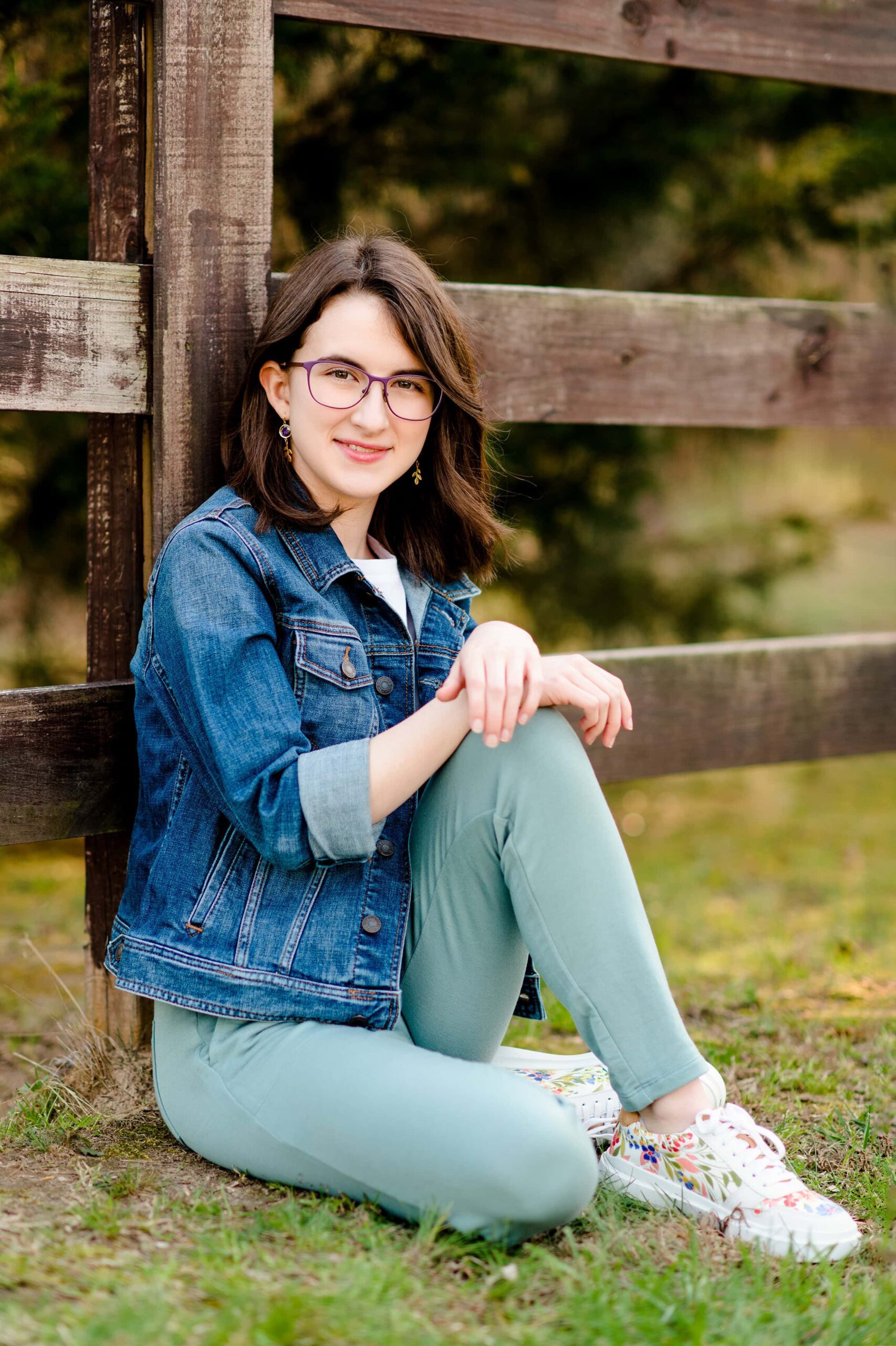 seated against a fenceline a senior poses with one leg up wearing a jean jacket, white shirt, and green pants with flower shoes