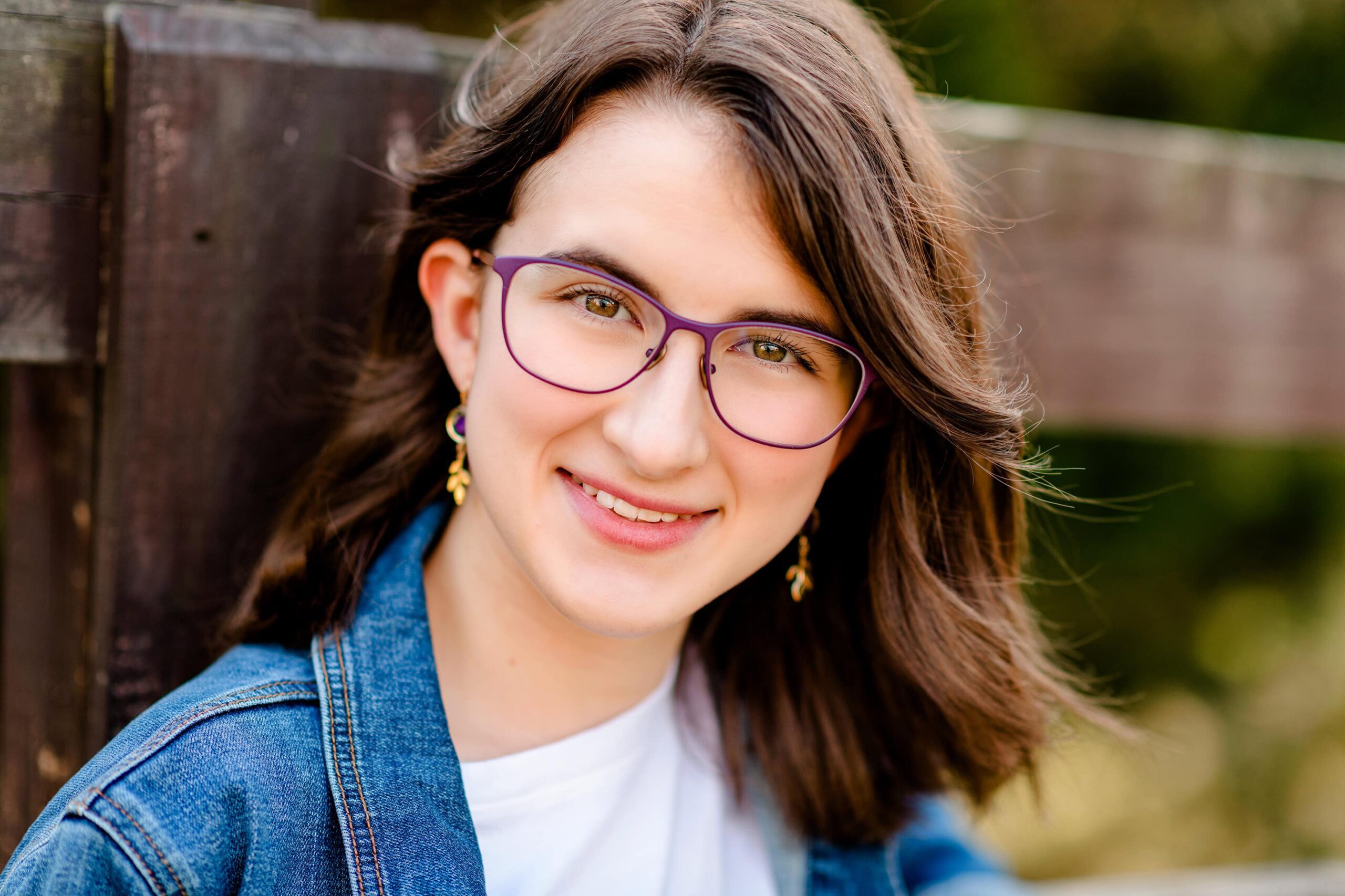 close up senior portrait of a dark hair grad with glasses wearing dangle earrings and a jean jacket