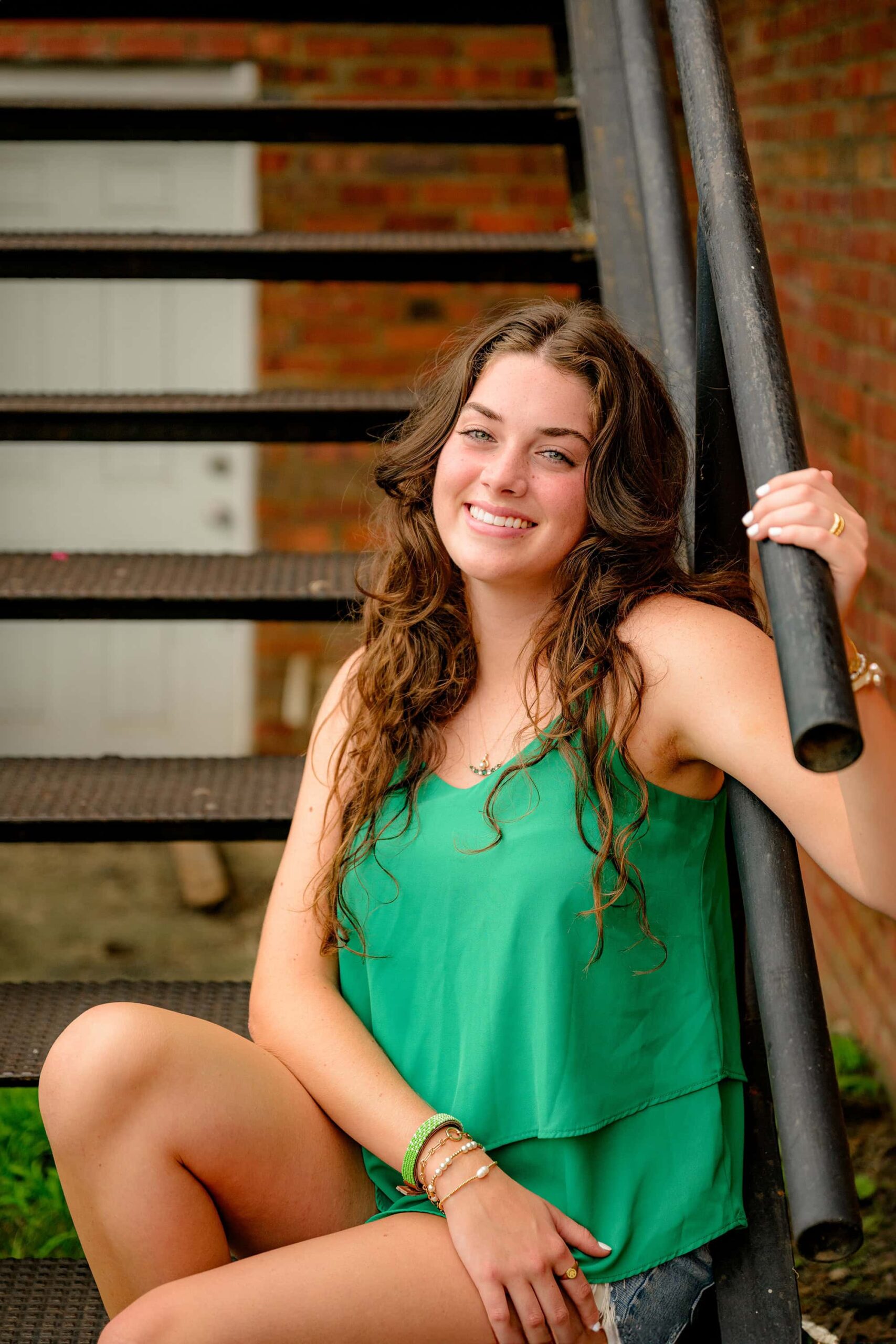 Emma Hines, a senior from Cuthbertson High School, sits on a metal staircase in downtown Waxhaw, holding onto the railing with a relaxed smile, wearing a green shirt, jean shorts, and matching green shoes.