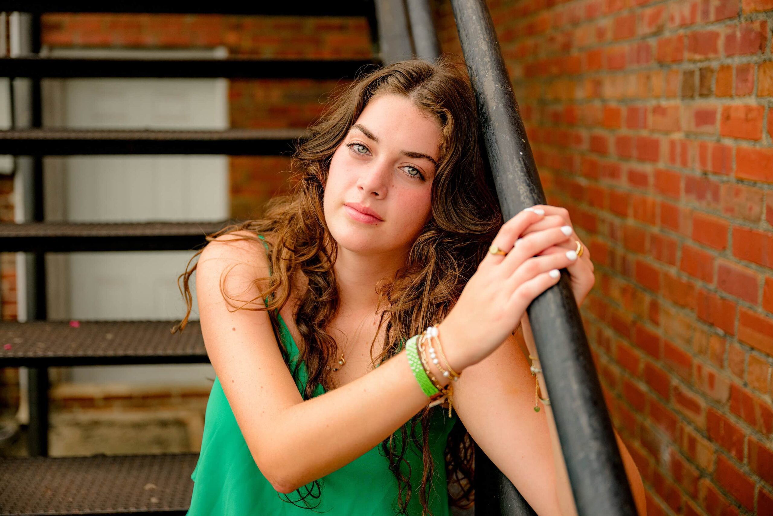 Leaning against an industrial metal railing, Emma Hines gazes into the camera, her striking eyes and dark hair standing out against the urban backdrop during her senior session in downtown Waxhaw.
