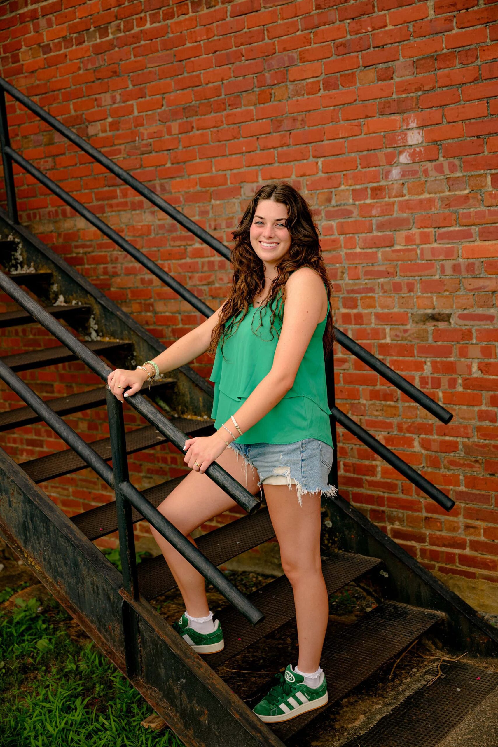 Emma Hines stands confidently on a staircase, each foot on a different step, showing off her green shoes in this full-body senior portrait taken in downtown Waxhaw.