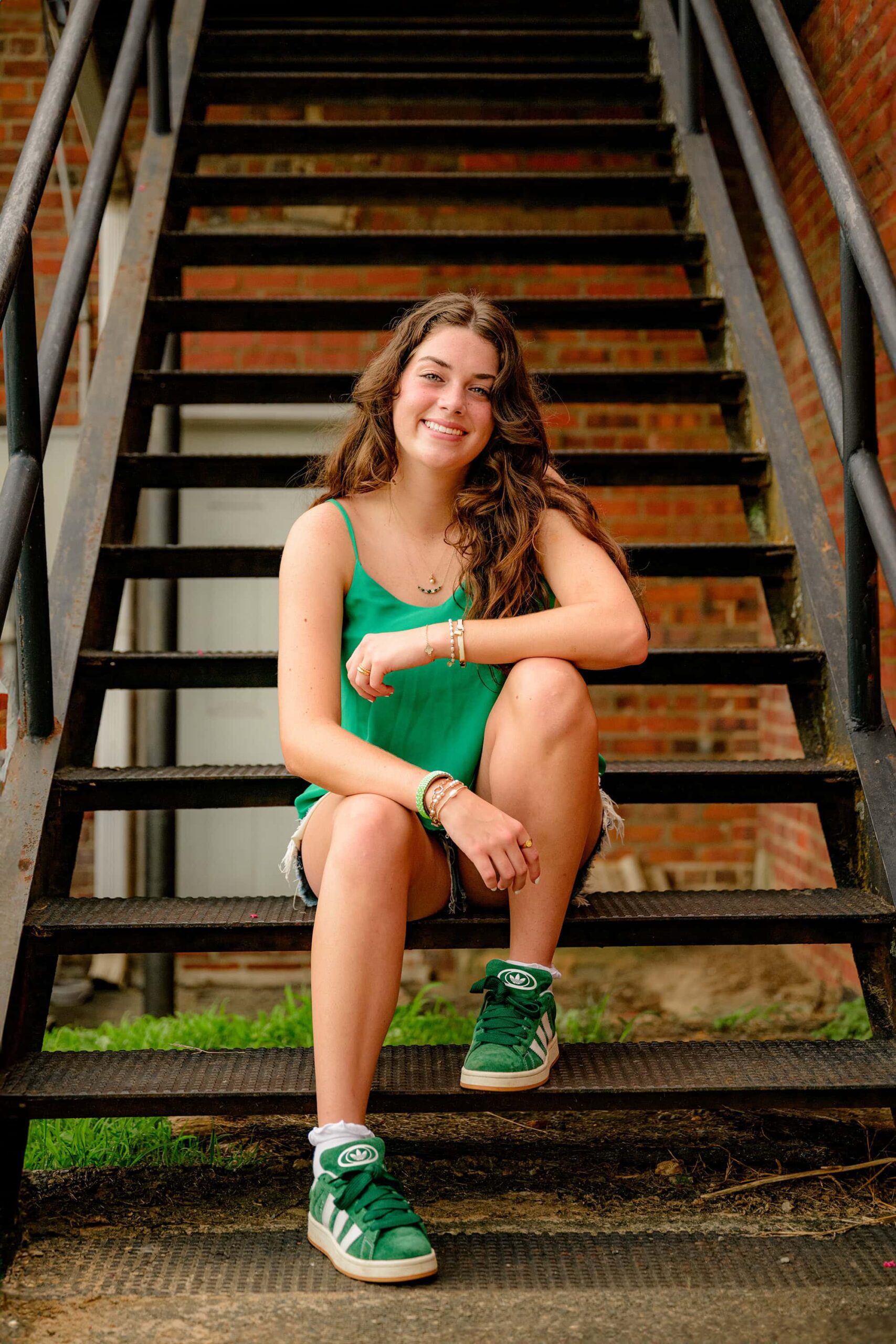 Sitting on a staircase, Emma Hines looks down toward the camera with a soft expression, one foot resting on a step and the other on the ground, her arms gently placed on her legs during her downtown Waxhaw senior session.