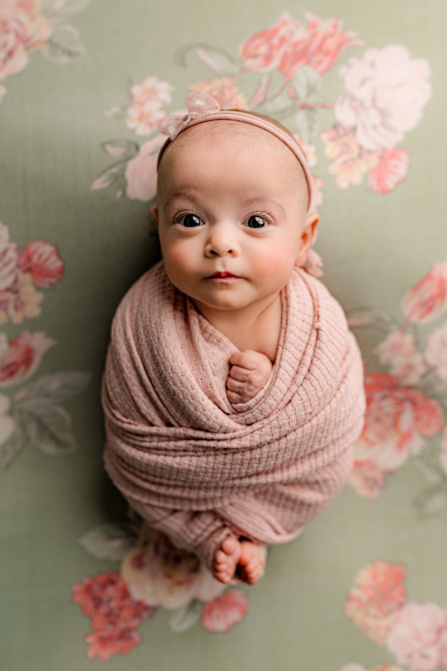 Awake newborn baby fully wrapped in a soft pink wrap, with her tiny toes peeking out, posed on her back on a green and pink floral background in the Waxhaw studio.