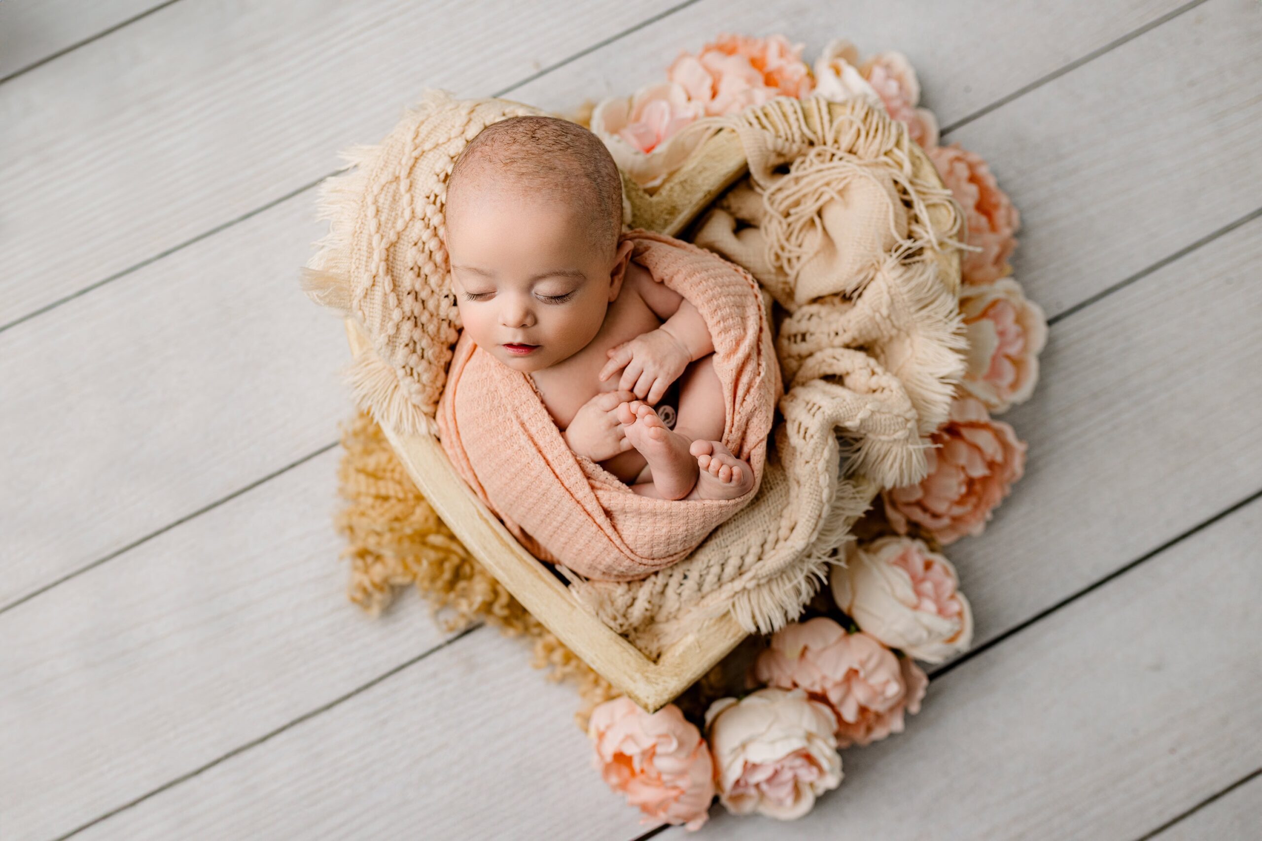 Newborn baby girl curled up in a heart-shaped prop, wrapped in a peach-colored wrap, resting on a wooden floor with peach flower accents surrounding her.