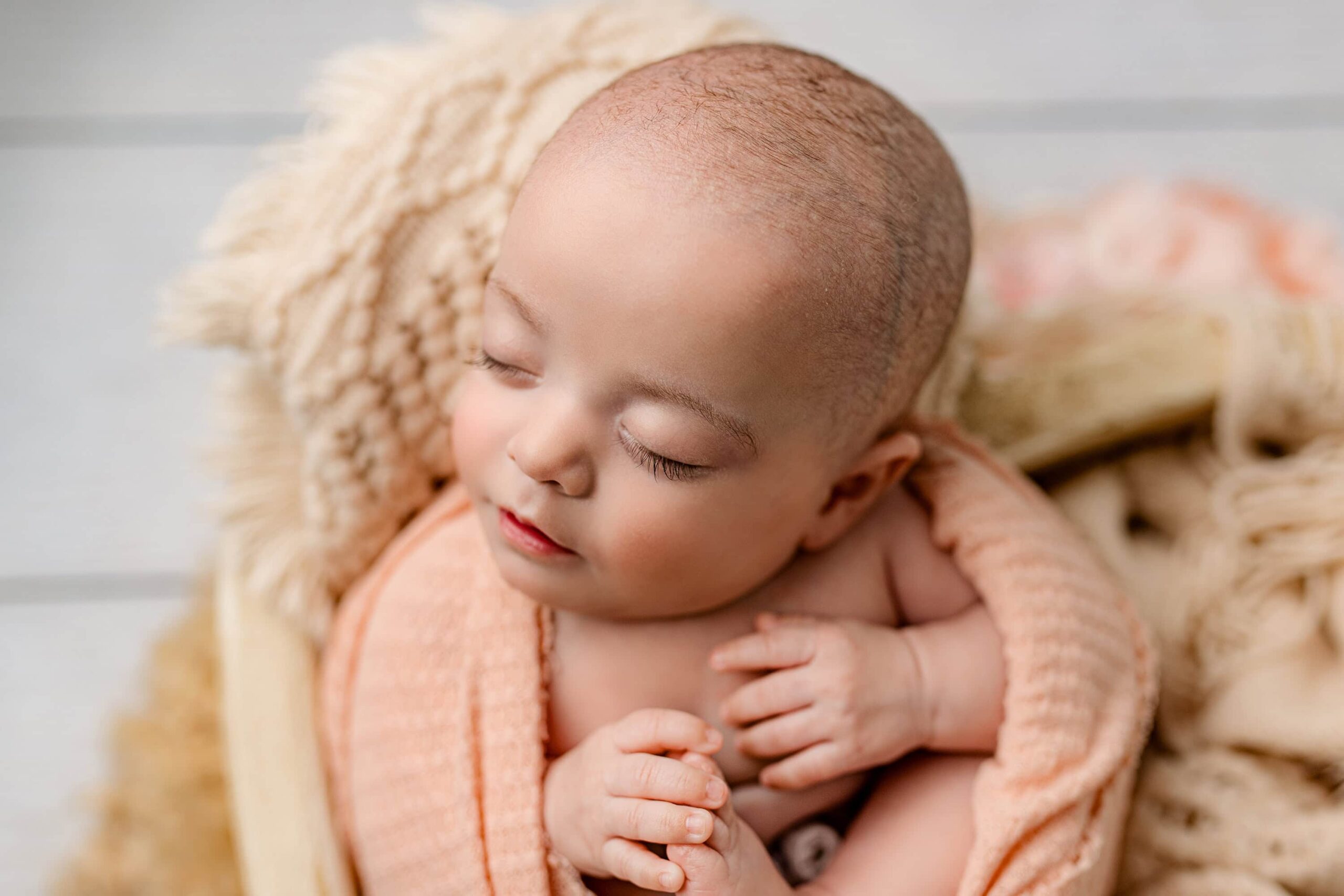 Side profile of a newborn baby girl wrapped in a peach-colored wrap, showcasing her long, delicate eyelashes against a soft peach-toned backdrop.