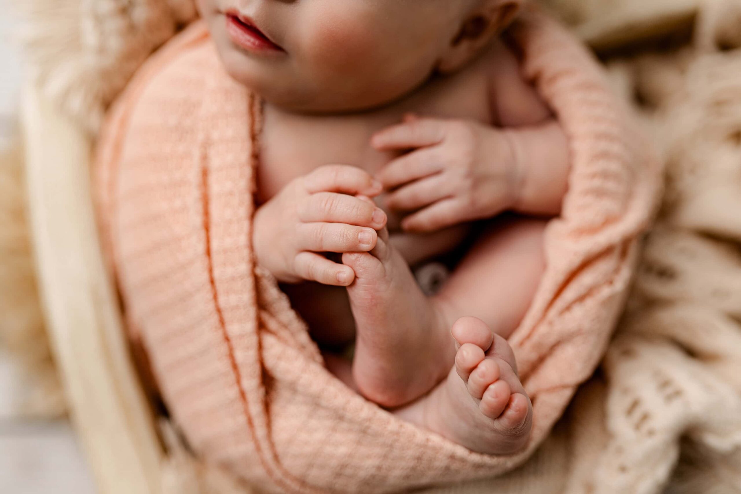 Close-up shot of a newborn baby girl's hands, feet, and legs, gently wrapped in a soft peach-colored wrap, with a peach-toned background.