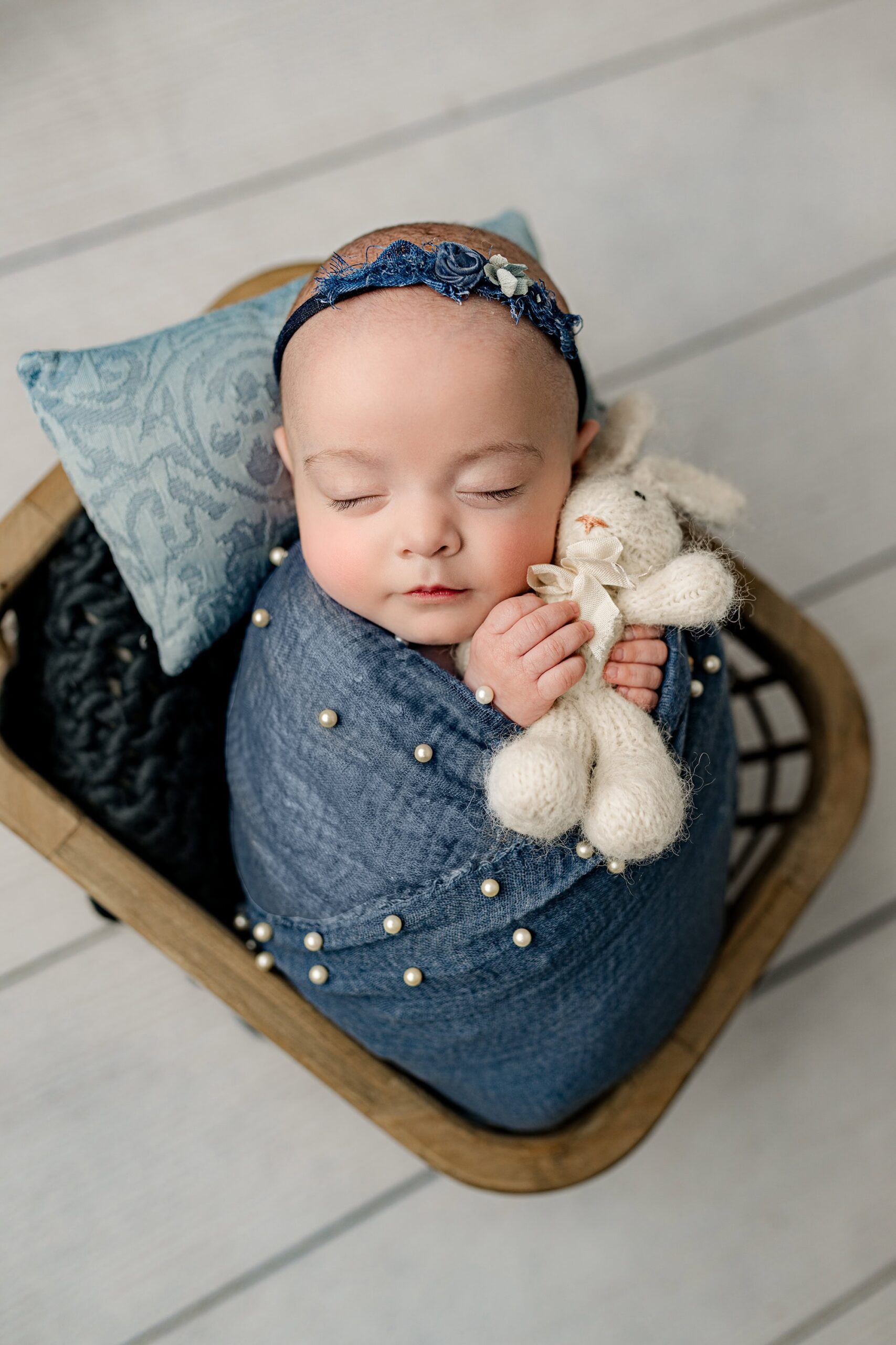 Asleep newborn baby girl, wrapped in blue with pearls, cuddling a white bunny in her cozy brown basket, with blue accents, captured in the Waxhaw studio.