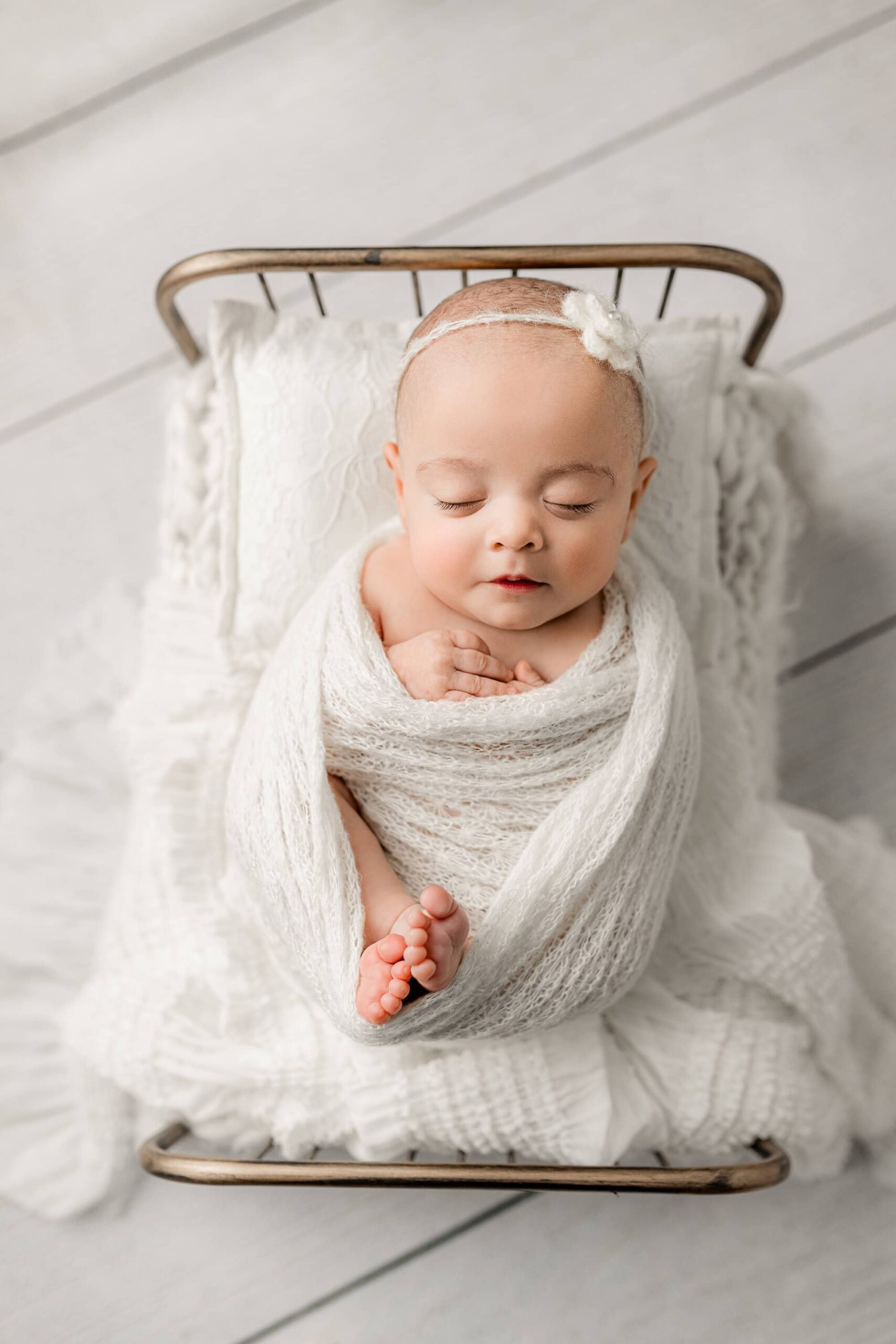 Newborn baby girl from Waxhaw, posed in a sheer white wrap and a pearl-accented white flower headpiece, lying on a gold-toned bed with her legs curled up.