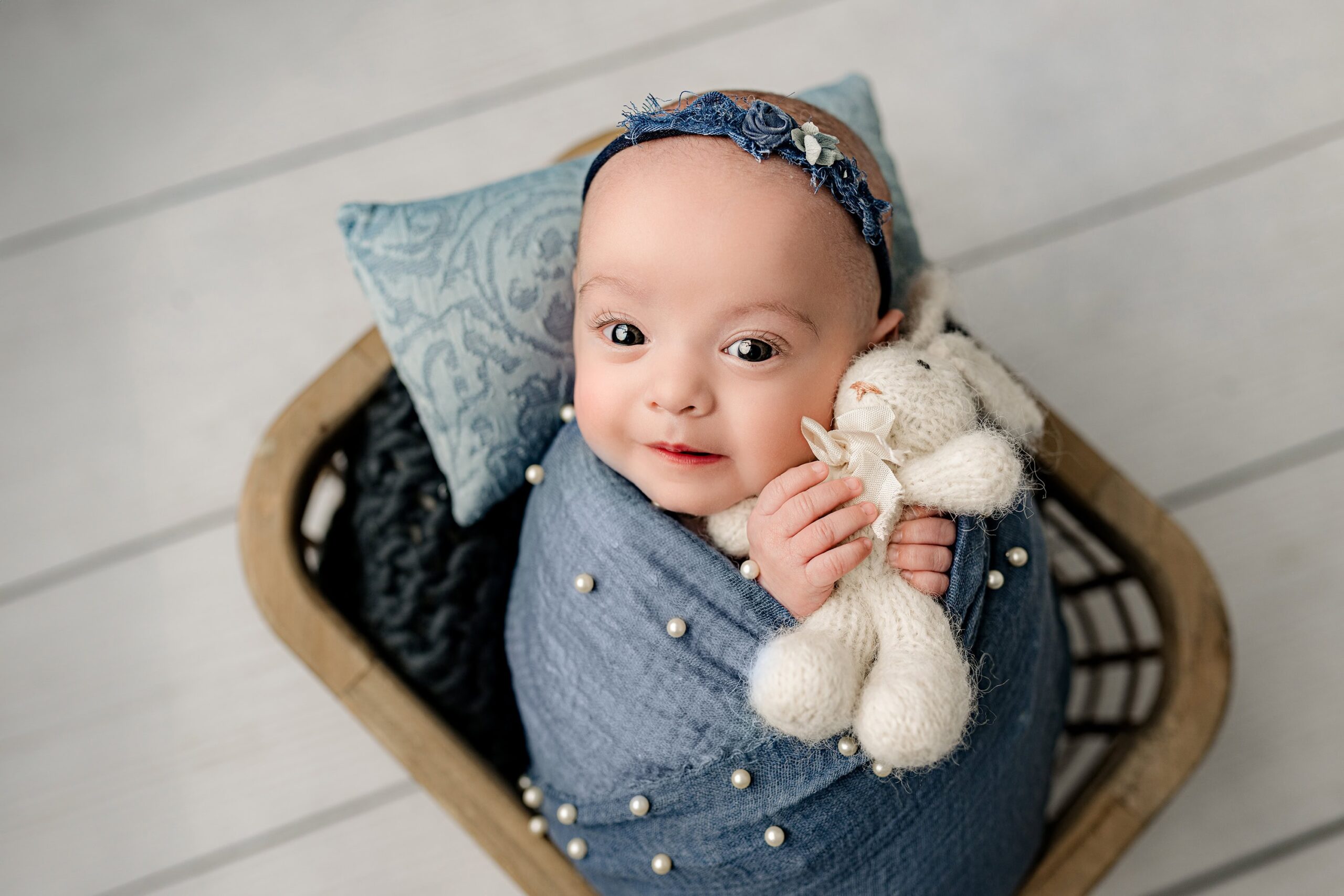 Awake newborn baby girl, wrapped in blue with pearls and wearing a flower headband, snuggling a white bunny posed on a blue pillow in the Waxhaw studio.