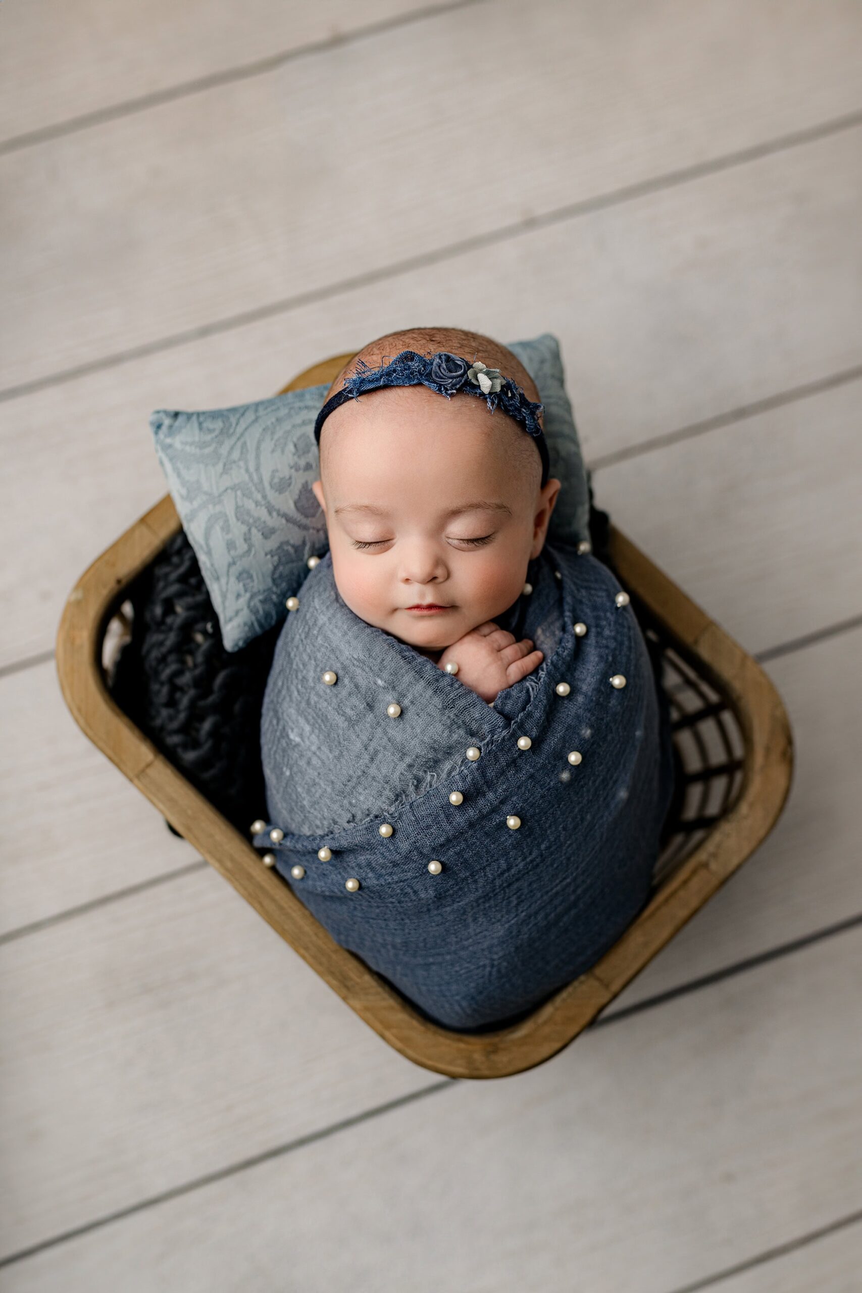 Newborn baby girl wrapped in a soft blue wrap with delicate pearls and a matching flower headband, peacefully asleep in a cozy basket in the Waxhaw studio.
