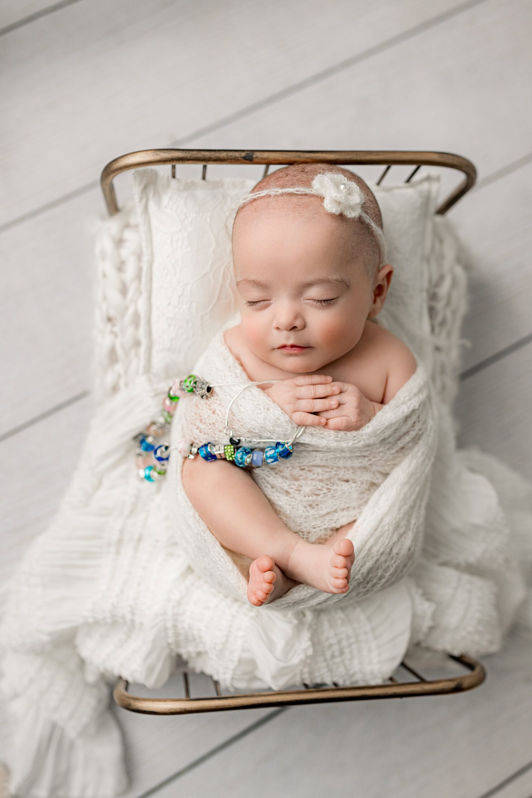Newborn baby girl resting on a gold-toned bed, holding her NICU necklace with milestone beads marking each milestone from her long NICU stay.