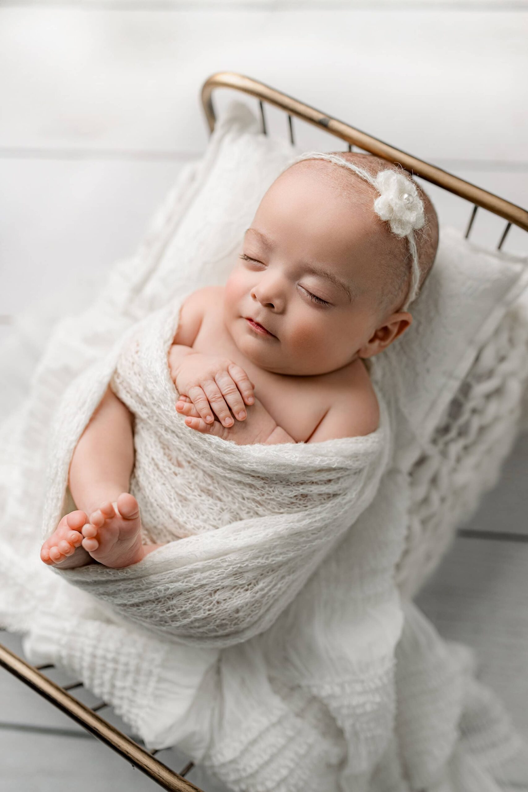 Side profile of a newborn baby girl in a sheer white wrap with a pearl-adorned flower headpiece, showing her hands peeking out of the wrap and curled legs.