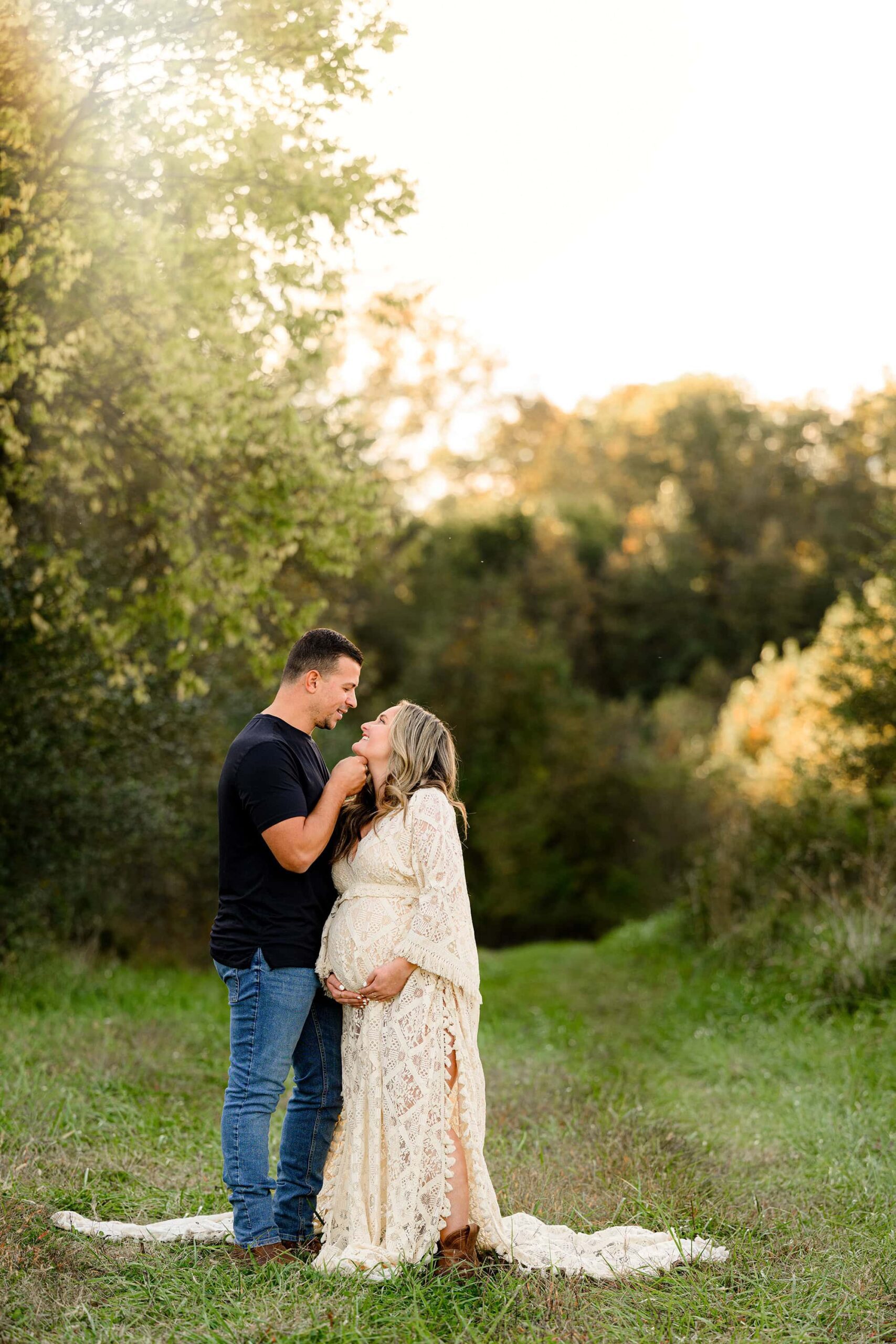 couple stands in an open grassy area in charlotte. dad is wearing a black shirt and jeans and has his hands under mom's chin. mom is wearing a cream colored boho style gown with her hands under her belly looking up at her husband