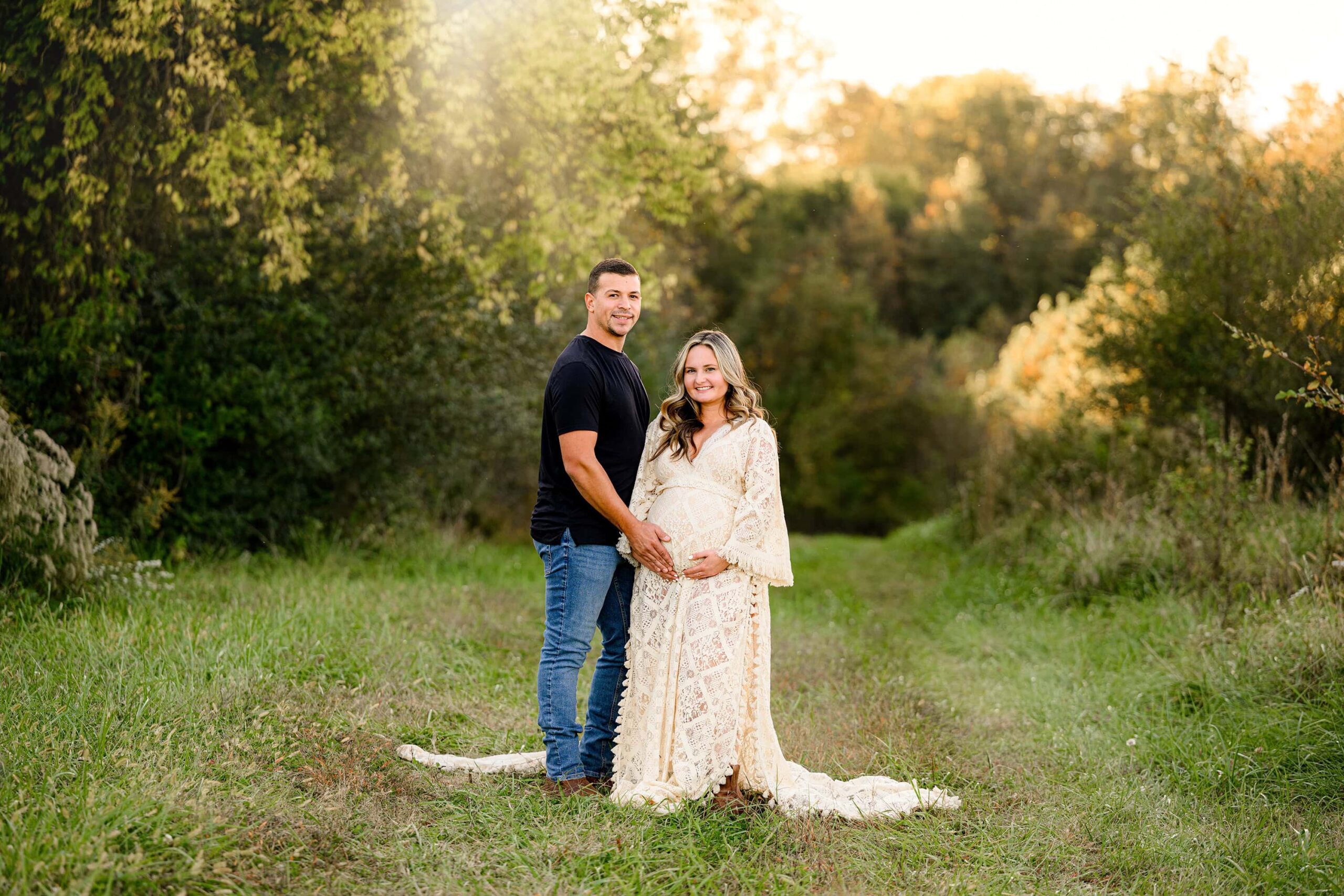couple stands together during their maternity session. Dad is dressed in a black shirt and jeans with boots and mom is wearing a cream boho style maternity dress and boots