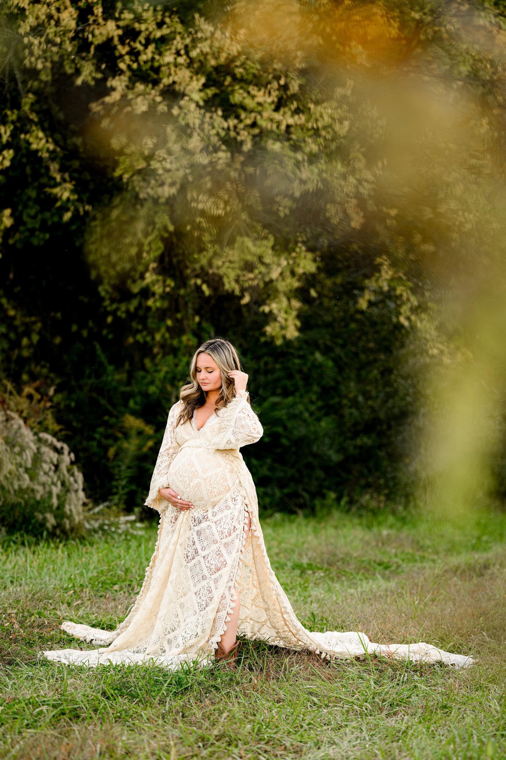 expectant mother from charlotte stands in a open grassy area wearing a boho style dress and cowboy boots holding her hair back and looking down at her stomach