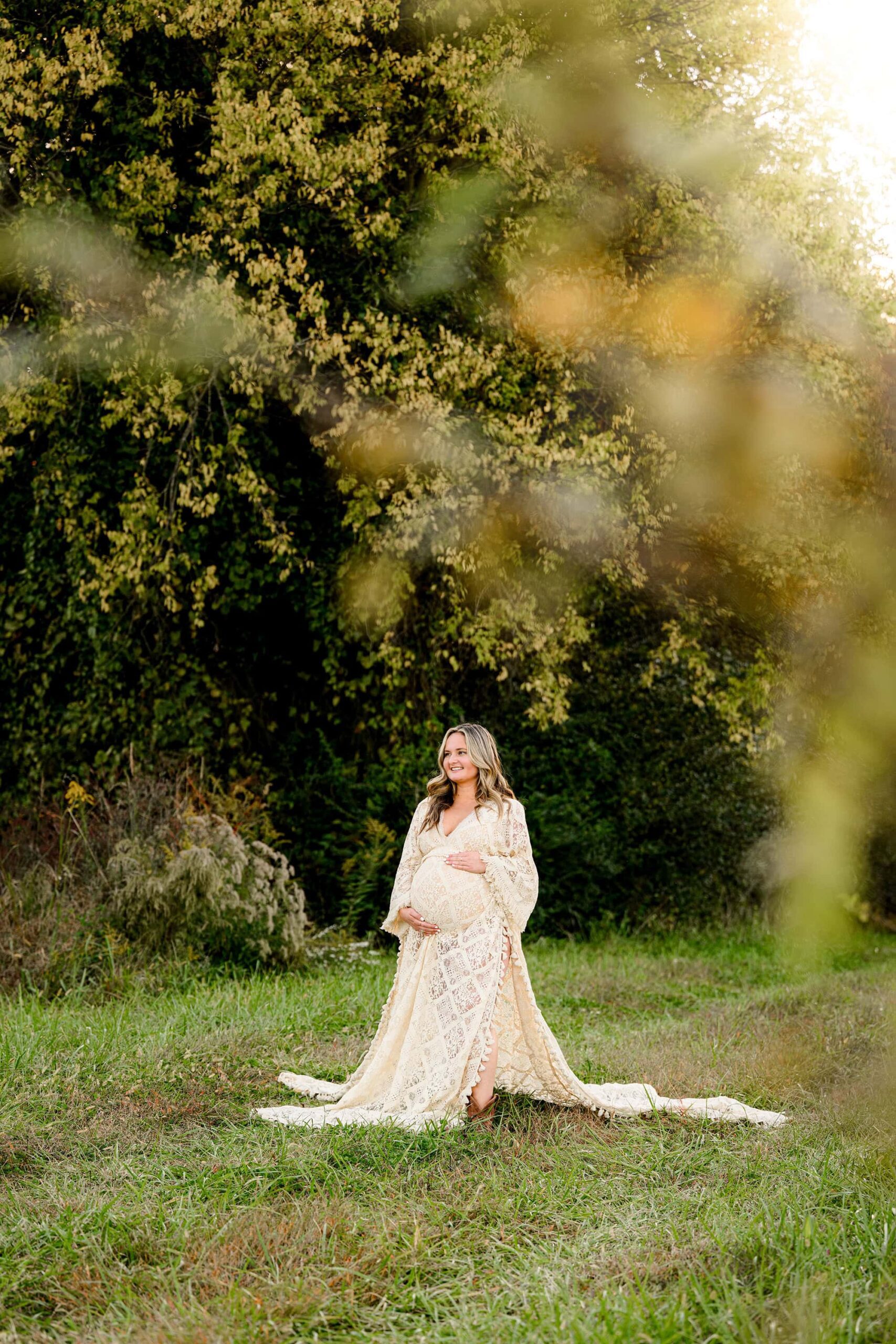 expectant mother to be stands in an open tree lined field wearing a cream lace boho style maternity dress with both her hands on her belly while she smiles