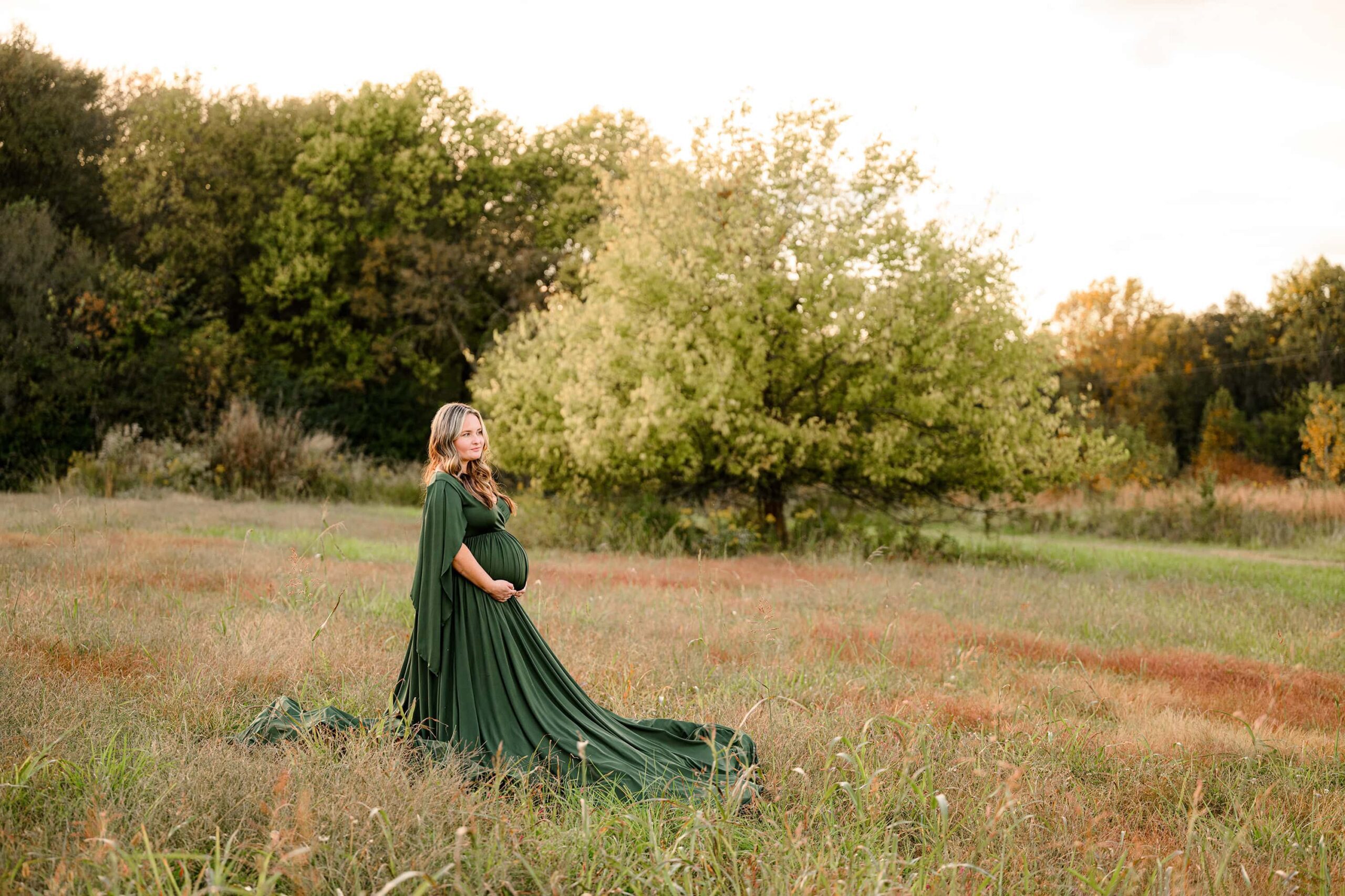 A mom to be stands in an open field in charlotte, nc dressed in a green dress with longer decorative sleeves. she is looking off in the distance and holding her hands under her belly