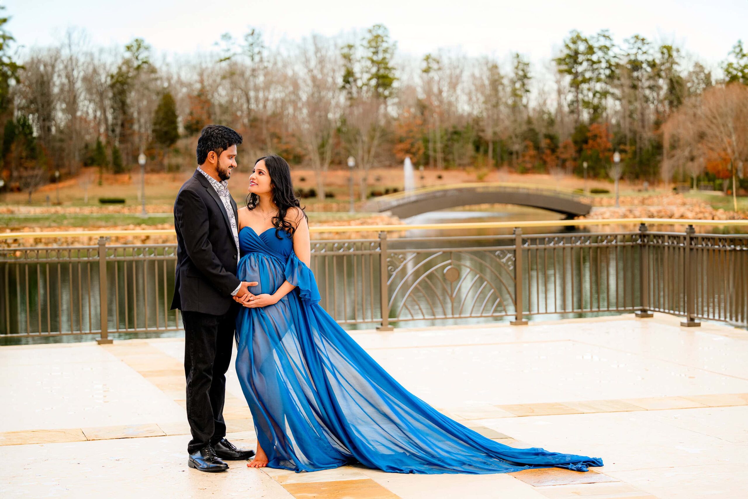 expectant couple stands together with a golden hour setting and a bridge in this charlotte area location. mom is wearing blue and dad is in black