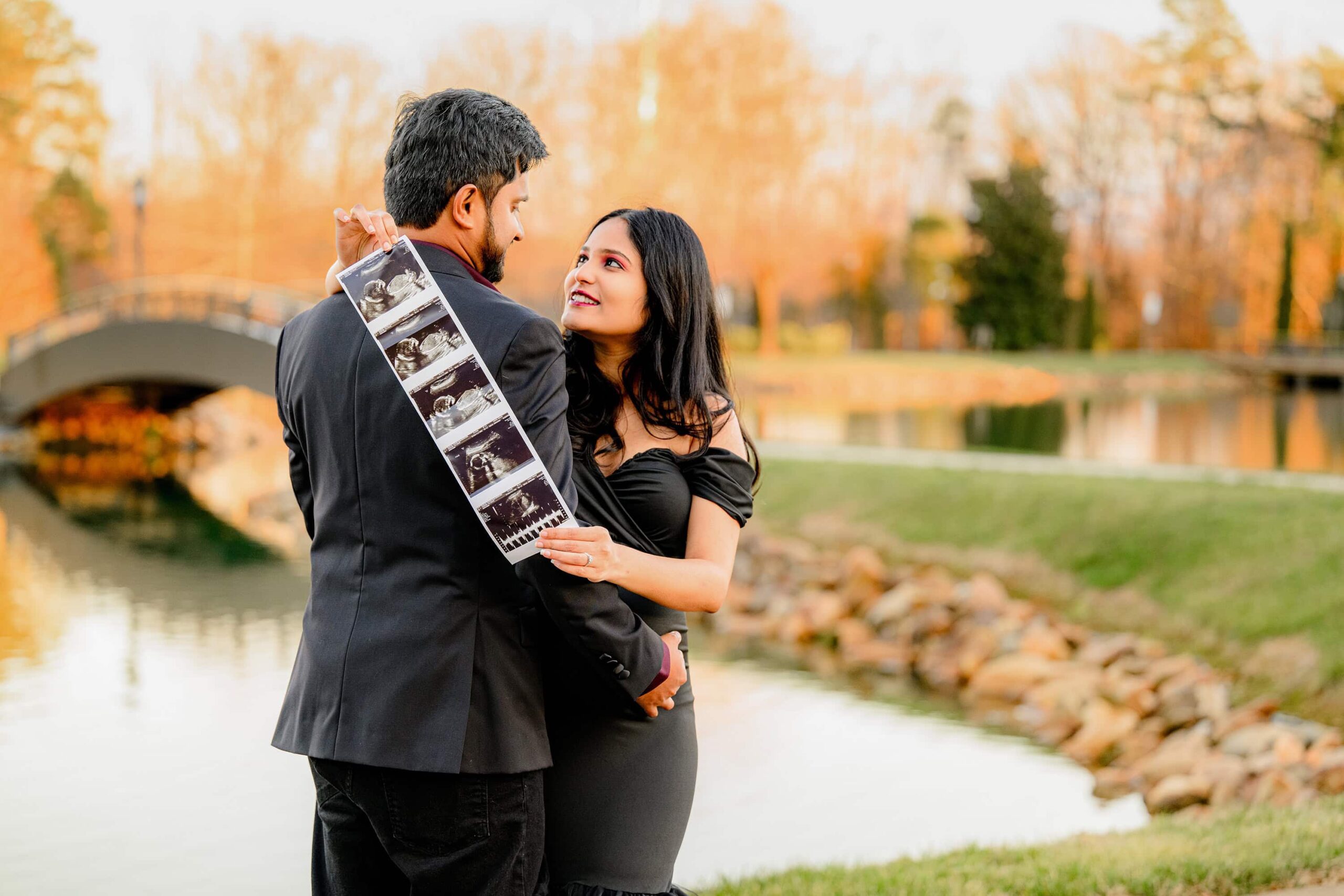 mother to be is posed holding an ultrasound photo while mom and dad embrace. mom and dad are both dressed in black