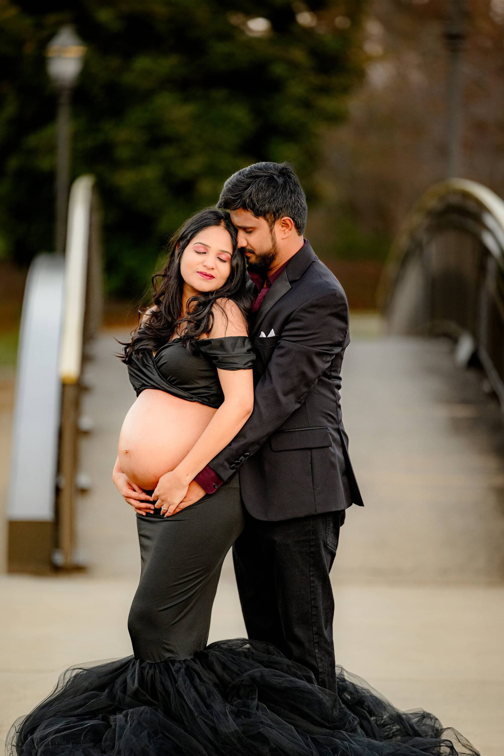 couple is posed together in a charlotte location with both of their heads leaning together and their hands on the bare belly of the expectant mom