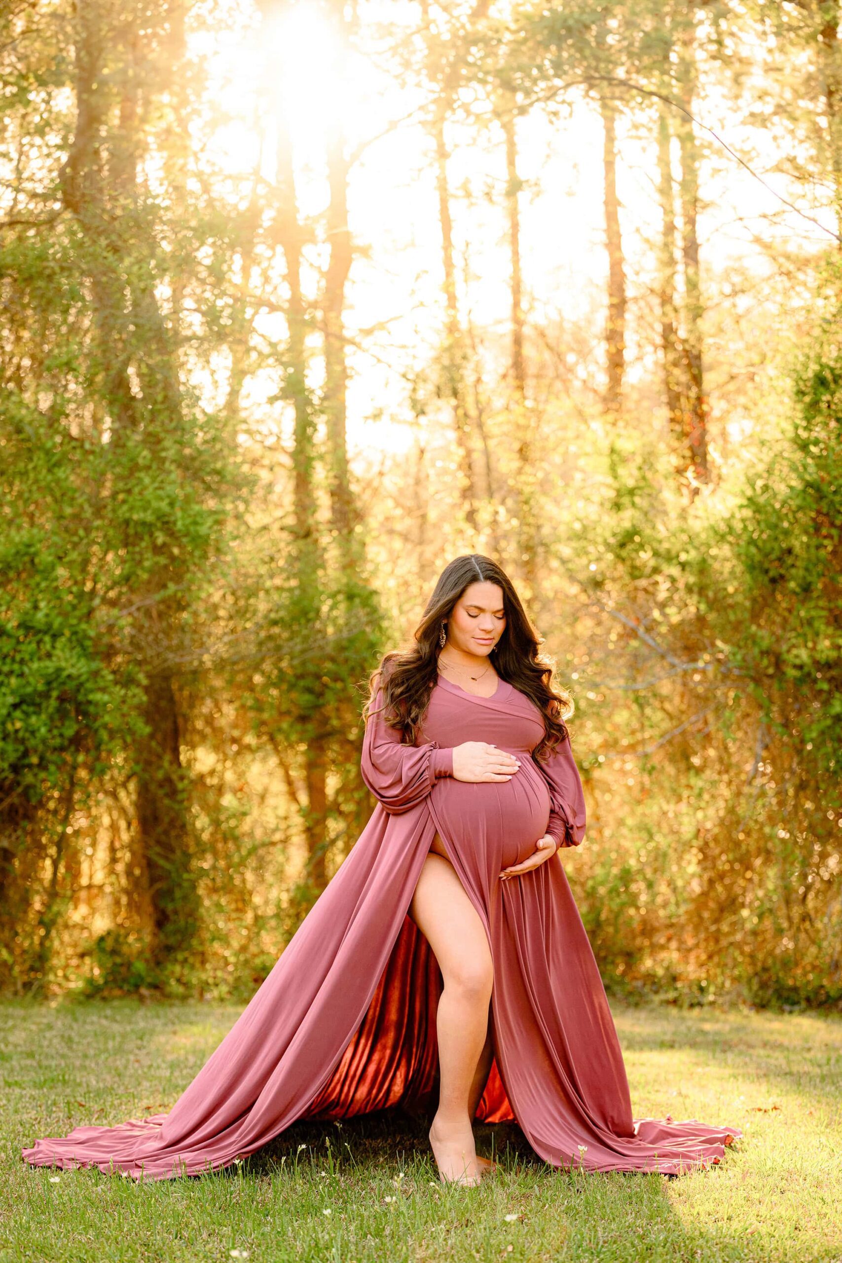 charlotte expectant mom poses with one leg out while wearing a rose pink dress with large slit and both hands on her bulging belly