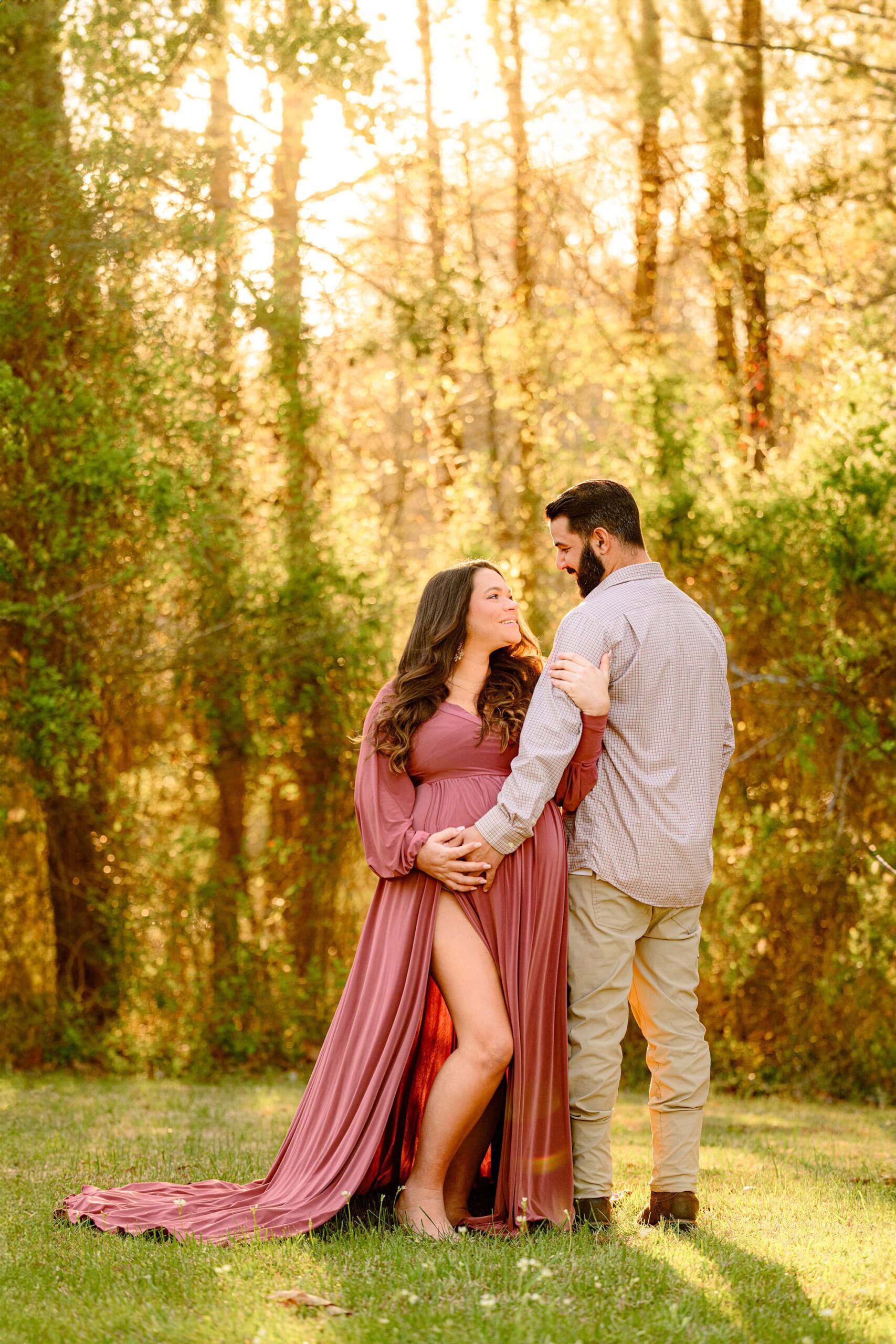 couple looks at each other with the spring golden light shining down on them during their maternity photoshoot. mom is wearing rose pink dress with a large pink slit and dad is wearing neutrals
