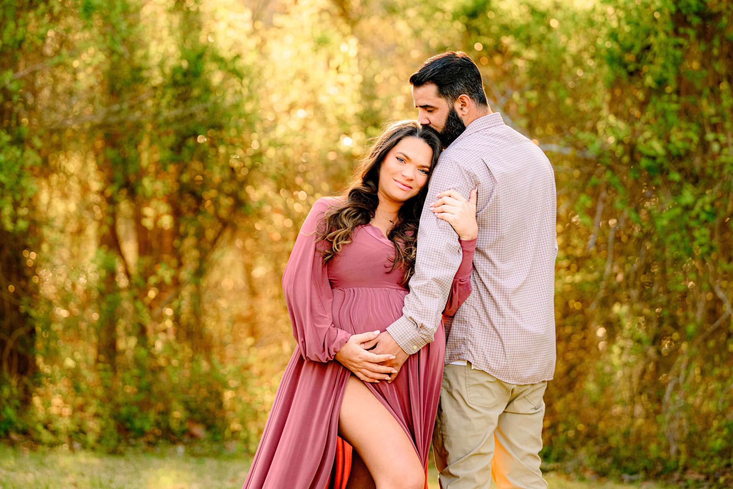 mom lays her head on dad's shoulder during her spring maternity photoshoot in a charlotte area park. mom is dressed in a rose pink dress