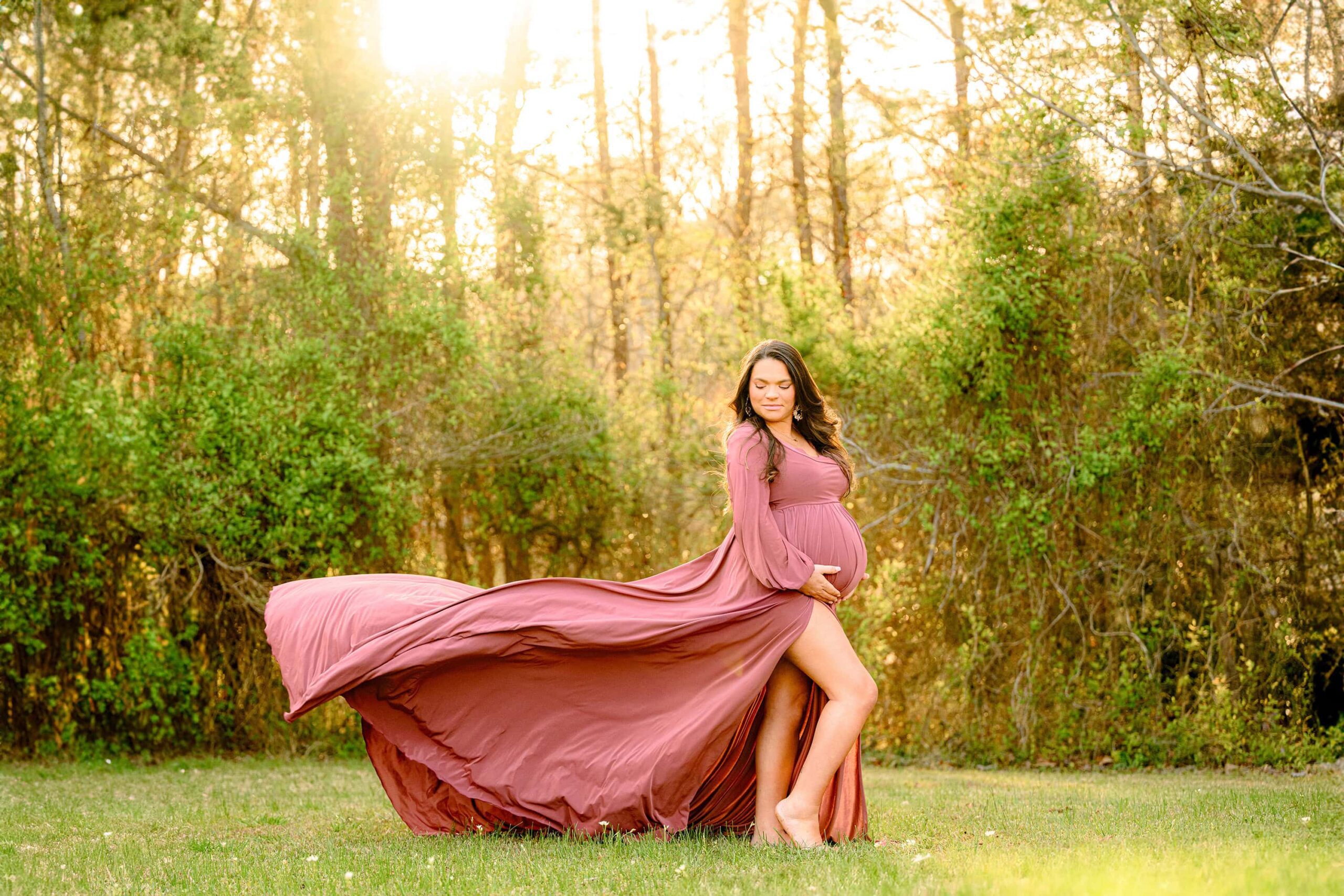 a dramatic dress toss photo of an expectant charlotte mother. the dress is rose pink with a large slit and she is posed holding her baby bump and looking down toward the ground
