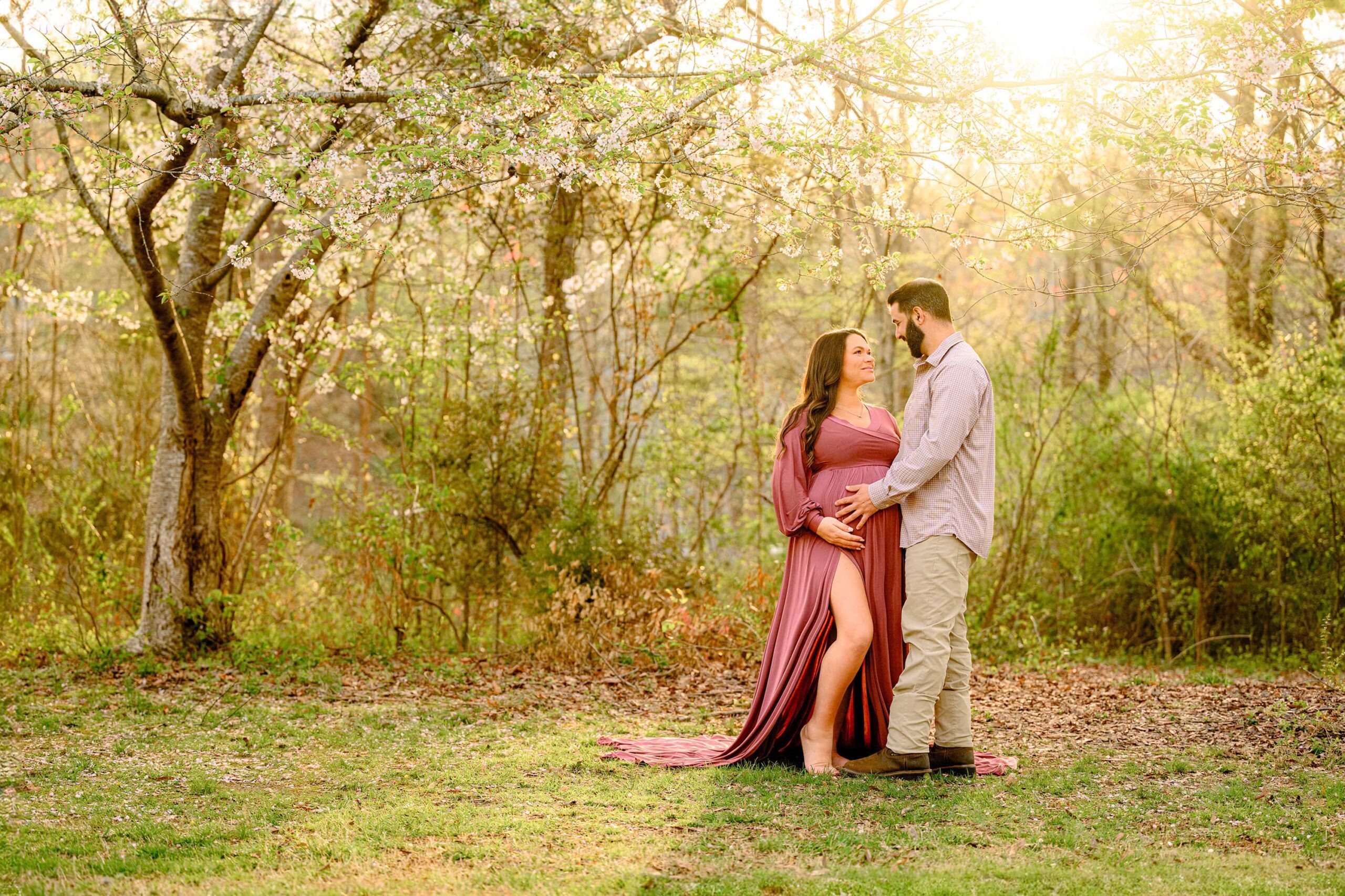 mom and dad are looking at each other with the sun shining down on them and the spring tree blooms. mom is wearing a rose pink dress with a large slit and dad is wearing brown and both have their hands on mom's belly