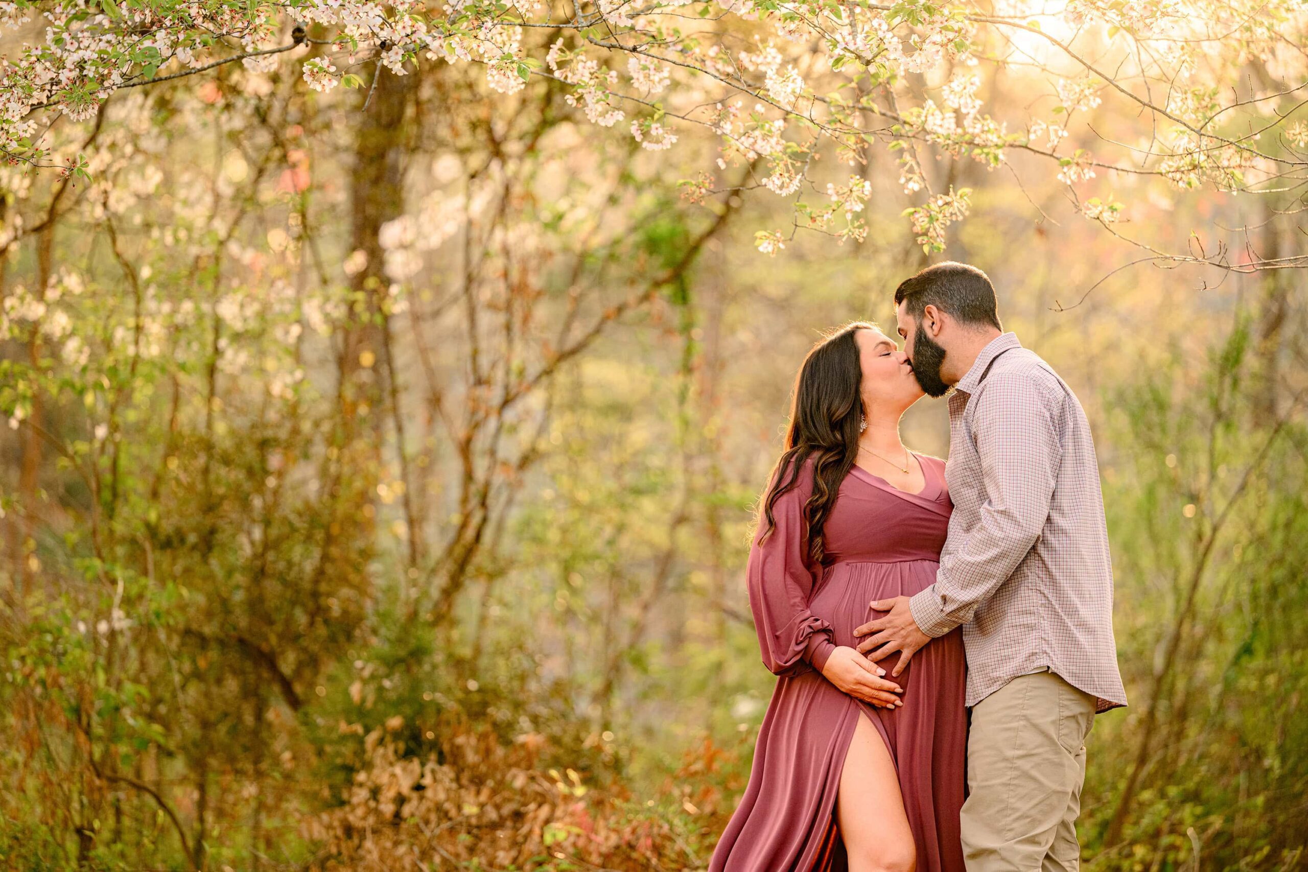 couple kisses under a spring tree during their charlotte maternity photoshoot. mom is wearing a rose pink dress and dad is wearing browns