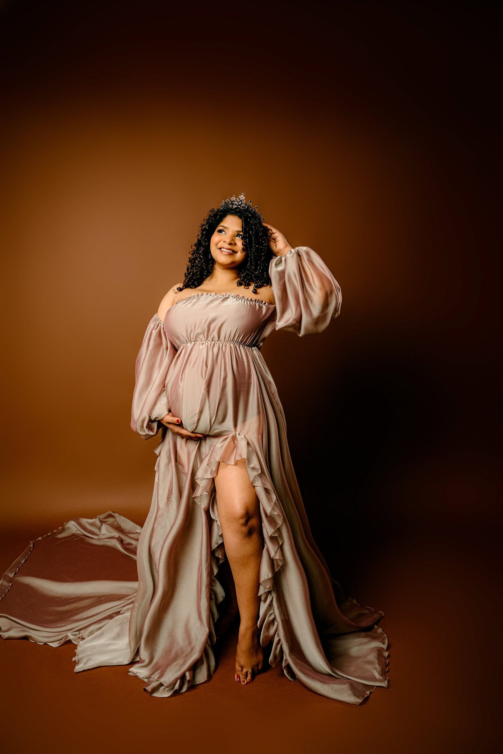 pregnant mother stands with one hand on her baby bump and one hand on a crown on her head. she is posed on a brown background and wearing a client closet brown maternity dress