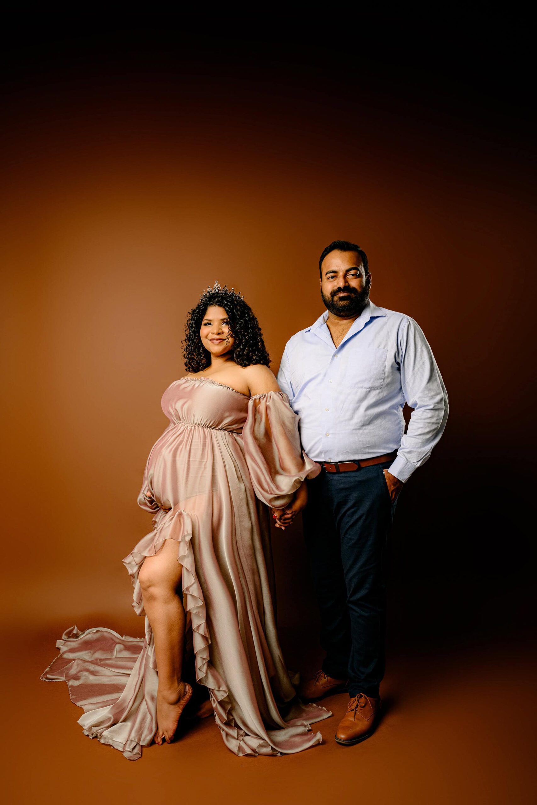 expectant couple poses together hand in hand with mom posed with her leg out and dad beside her. they are standing on a brown background in a waxhaw studio. dad is wearing. a blue shirt and pants and mom is wearing a brown dress