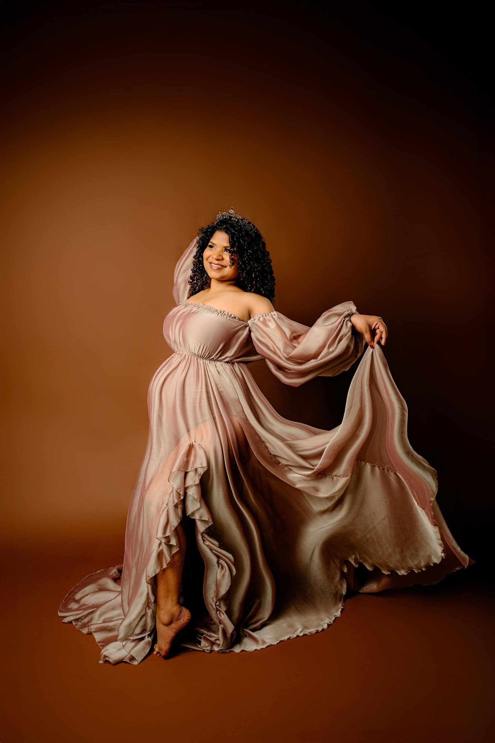 expectant mother with dark curly hair and wearing a brown dress holding the edge of her dress and a crown on her head