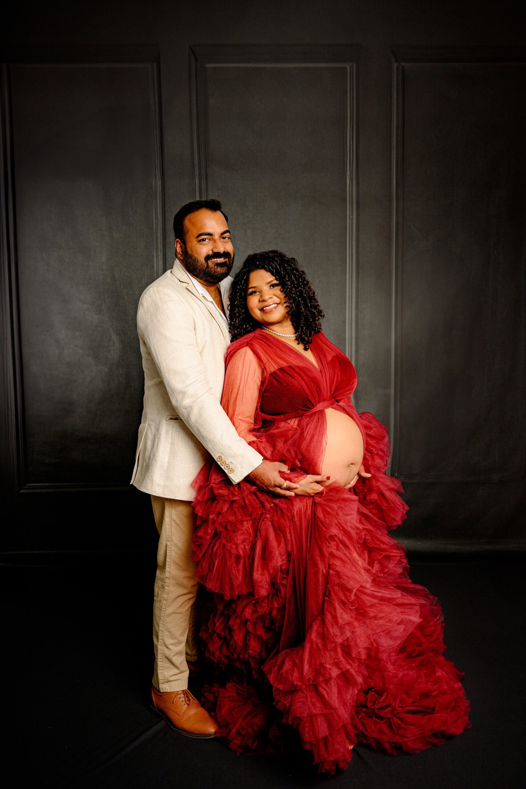 expectant couple stands together with arms around each other and hands on mom's belly. maternity mom is wearing are red tulle dress and dad is wearing a neutral jacket and pants