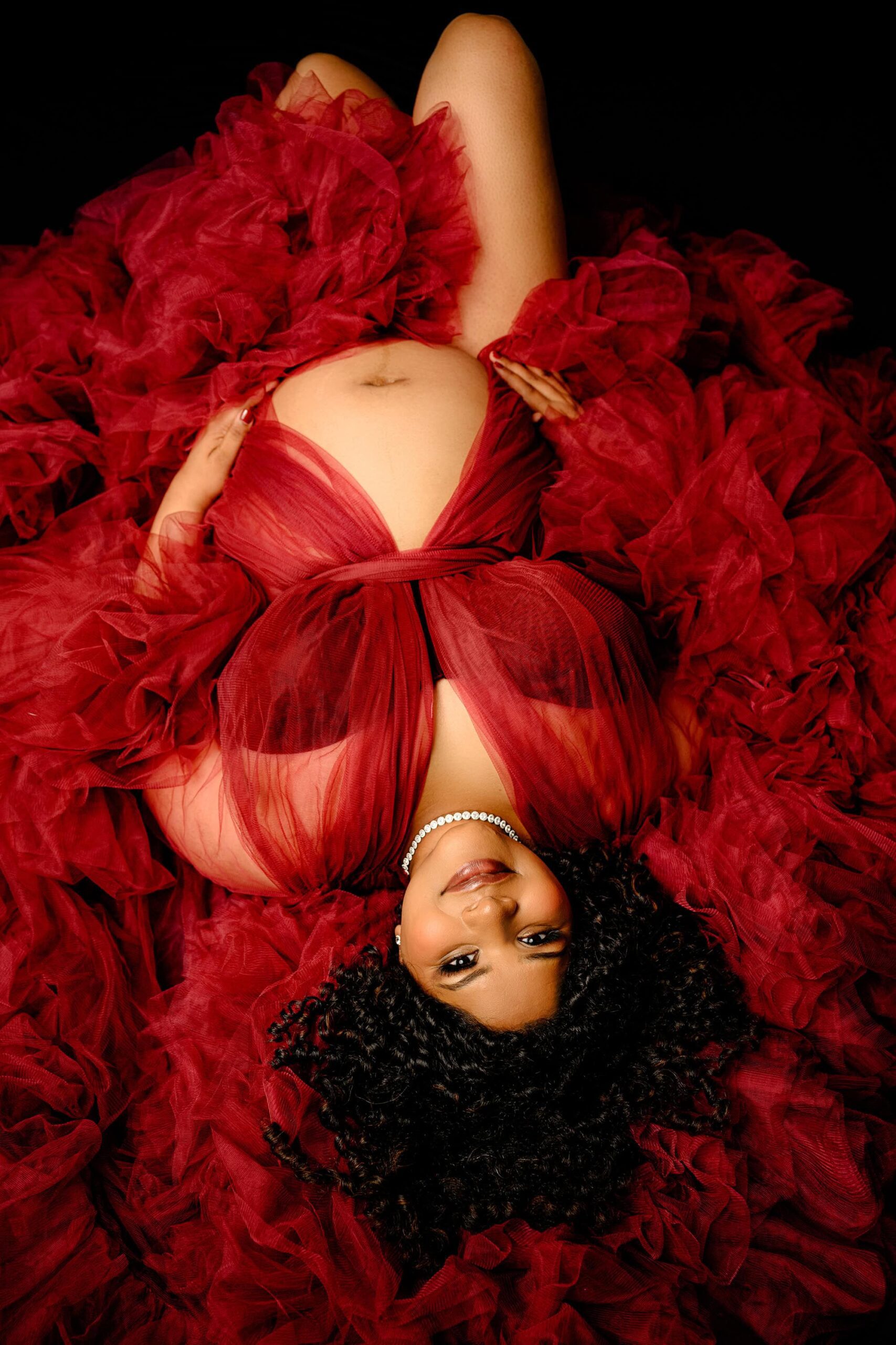a pregnant mother lays down with her expectant belly exposed and her red tulle dress all around her