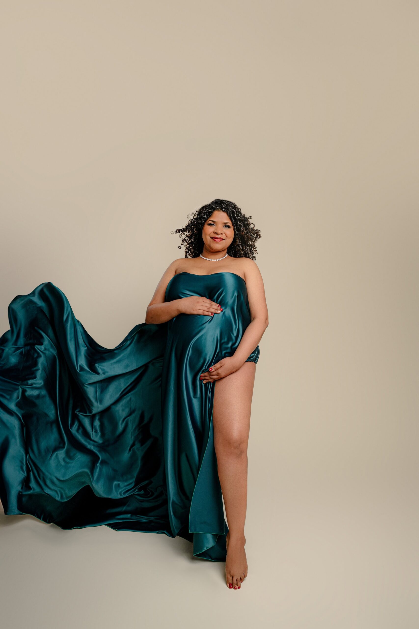 a green silk is billowing around a expectant mom as she is posed in a dramatic way with the silk covering her baby belly and her leg out