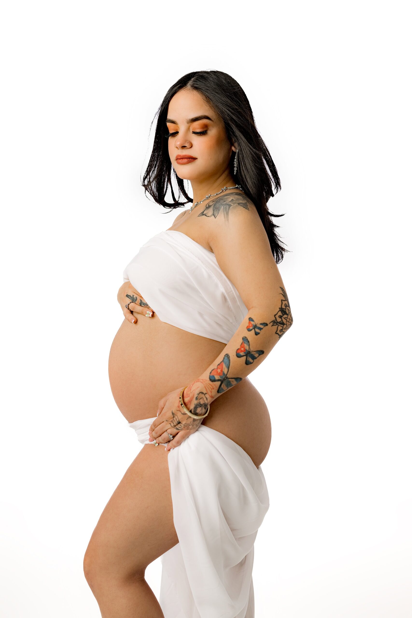 A timeless maternity image featuring soft white chiffon wrapped around the mother-to-be, showcasing her belly as she gazes downward, revealing her tattoos.