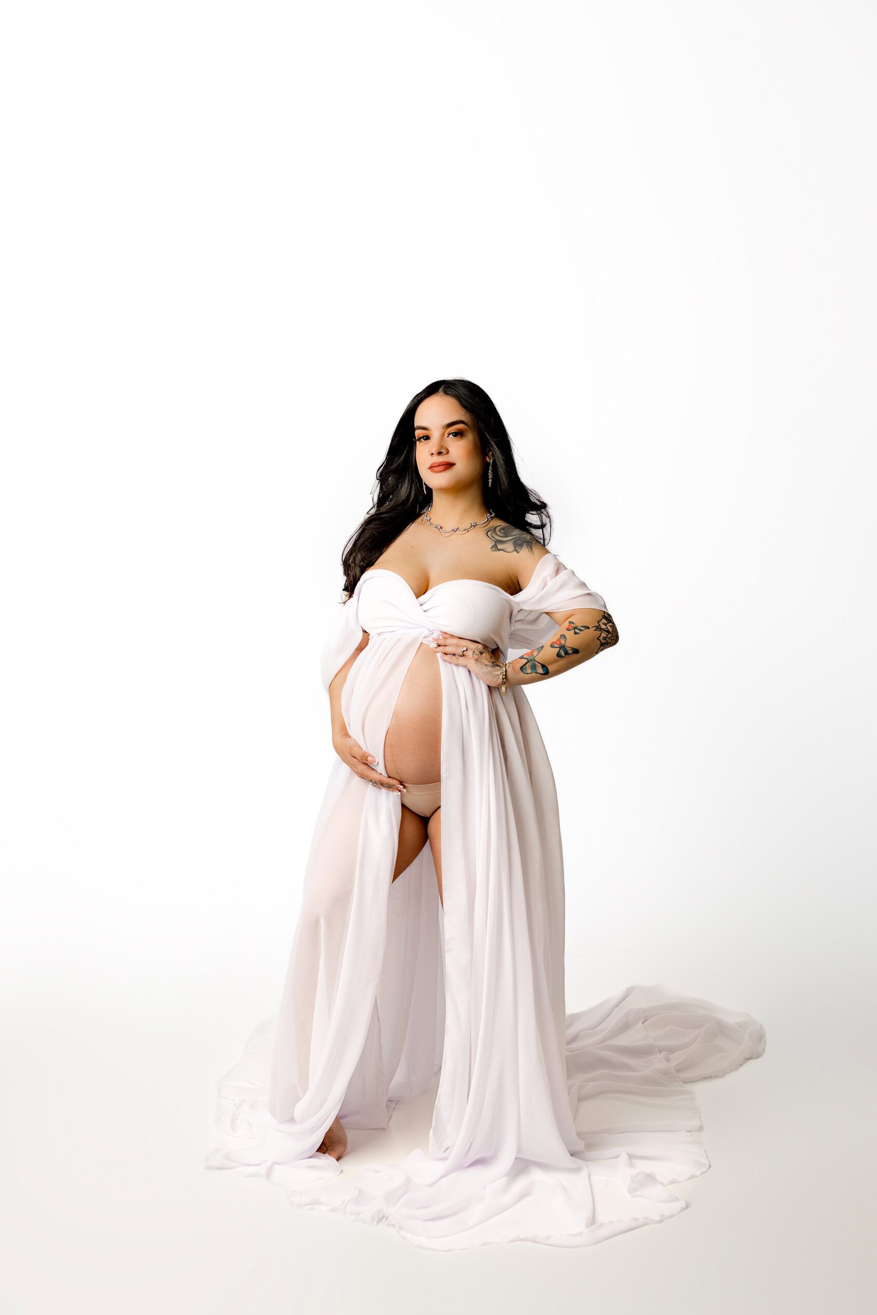Waxhaw Studio Session: A dramatic portrait of a dark-haired mom-to-be wearing a flowing white dress with a draped sleeve. The dress is gently opened, showcasing her silhouette as she gazes intensely at the camera. Her hands rest on her face, while her hair cascades down her back, adding an air of elegance and anticipation. The shot is captured with soft lighting to highlight the details of the dress and her serene yet powerful pose.