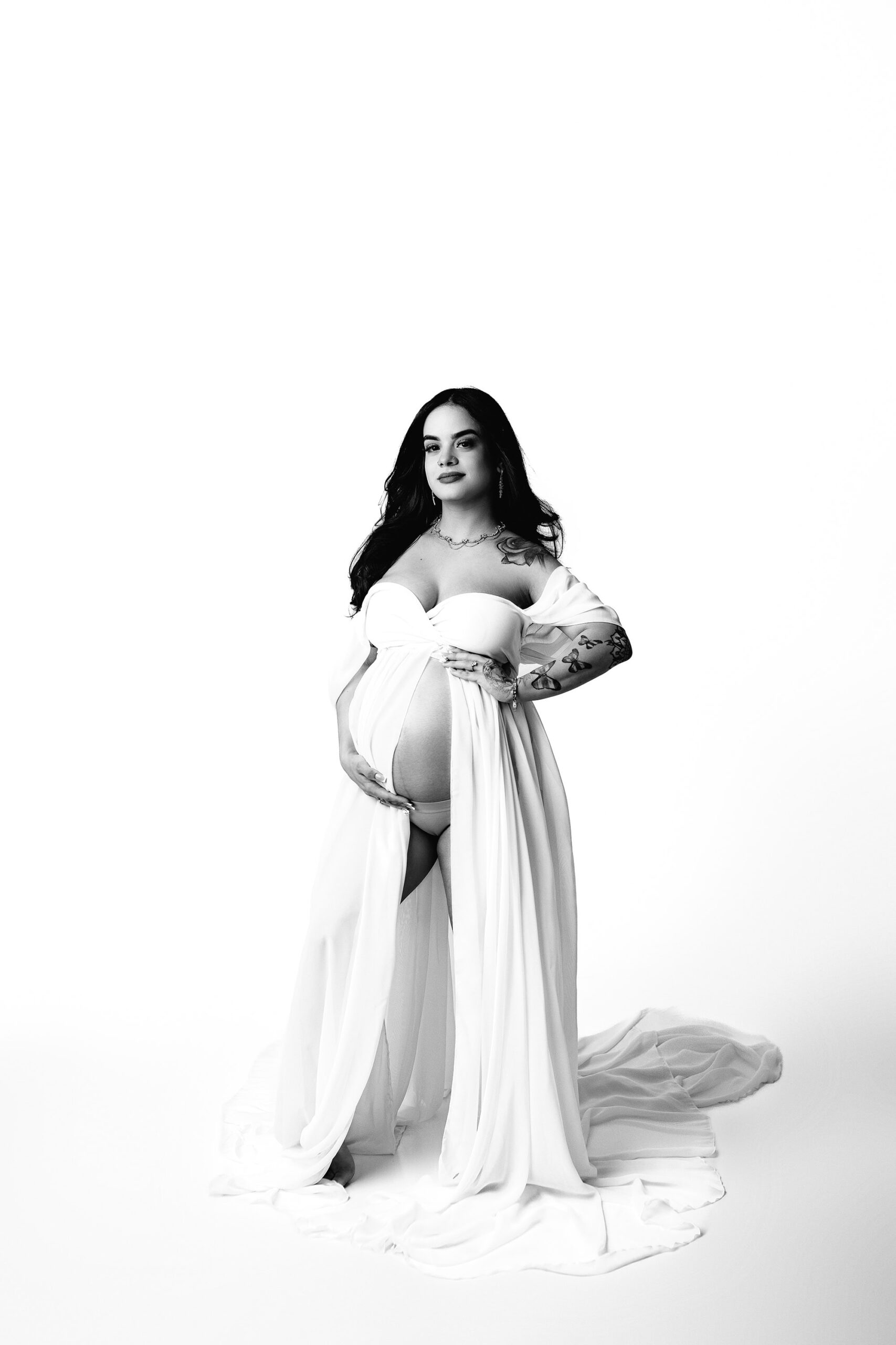 Charlotte, NC Area Mom-to-Be: A striking image of a dark-haired expectant mother, dressed in a white gown with a draped sleeve, gently parted to reveal a graceful silhouette. With her hands resting on her face and her gaze directed at the camera, she exudes a sense of calm strength. Her flowing hair adds to the beauty of the moment, creating a timeless feel in this intimate maternity portrait. The black-and-white version of the image adds depth and emotion to this delicate capture.