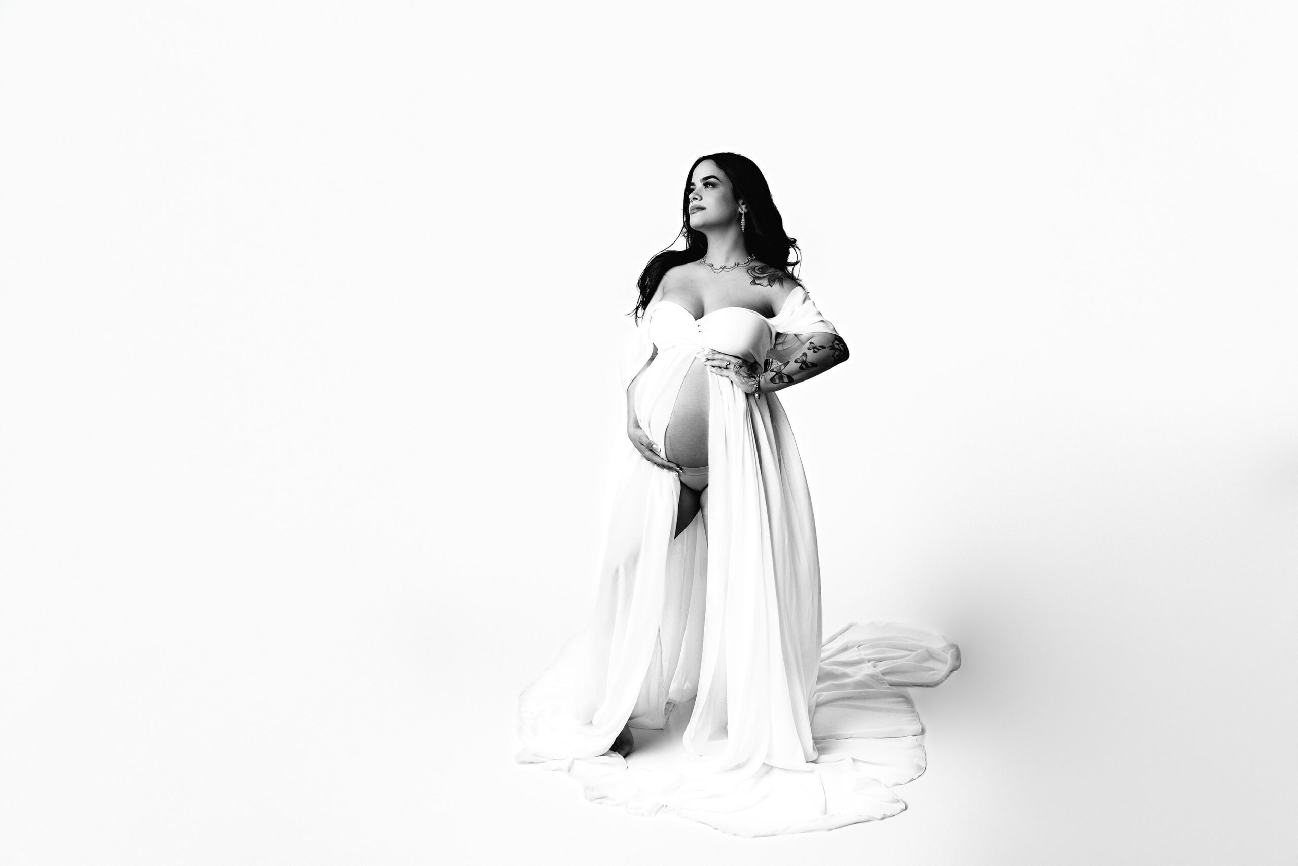 Black and white full-body image of a Charlotte area expectant mother in a dramatic pose, her belly showing as she cradles it with both hands, one leg extended with her toe popped.