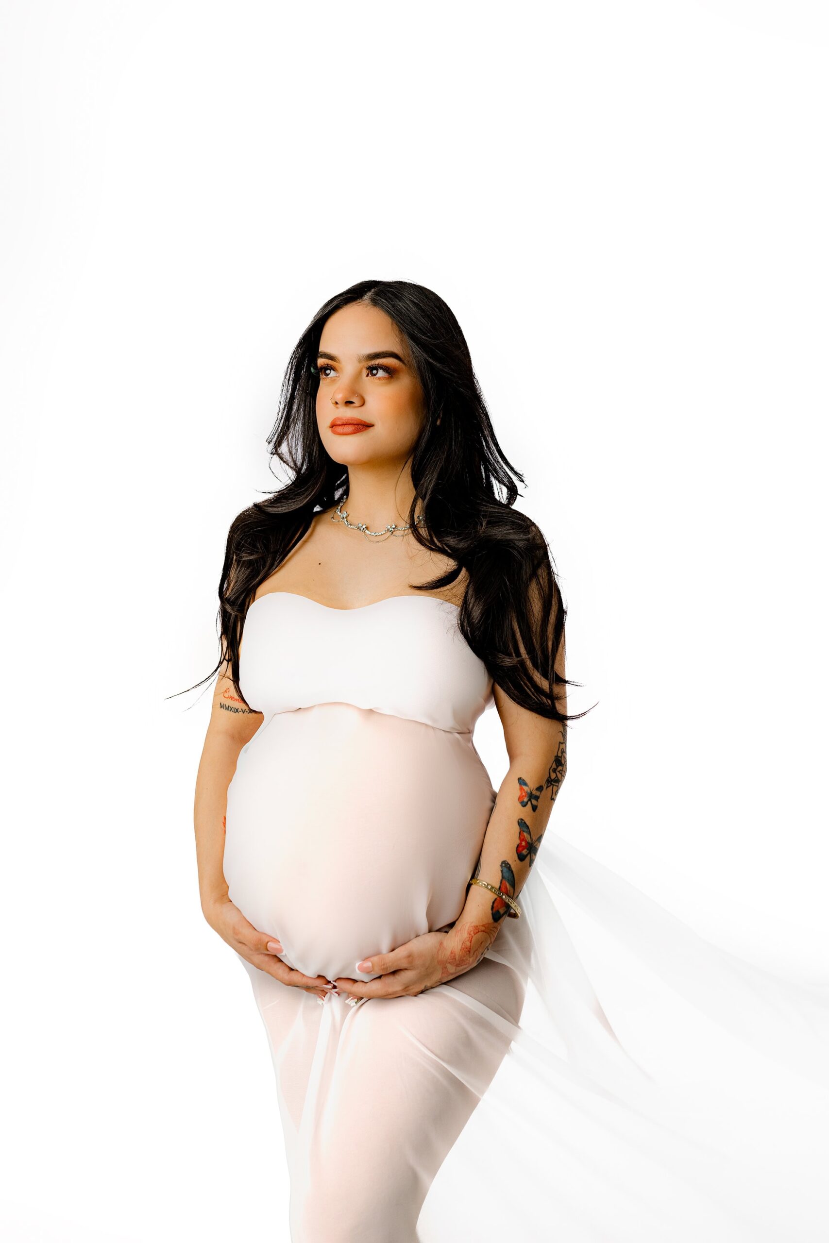 Elegant maternity portrait of an expectant mother from Charlotte, NC, with long dark hair and professional makeup, draped in white chiffon to highlight her bump.